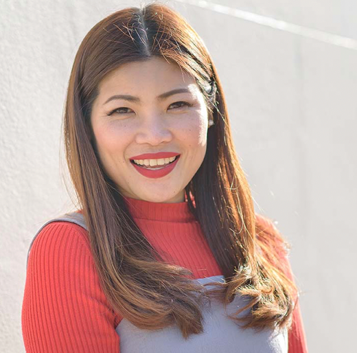 A woman with long brown hair smiling, wearing a red long-sleeve top and gray apron, outdoors in bright sunlight.