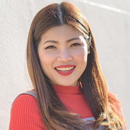 A woman with long brown hair smiling, wearing a red long-sleeve top and gray apron, outdoors in bright sunlight.