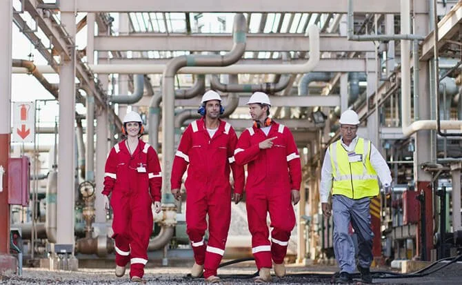Group of industrial workers in hard hats and red coveralls walking through a facility with pipelines.