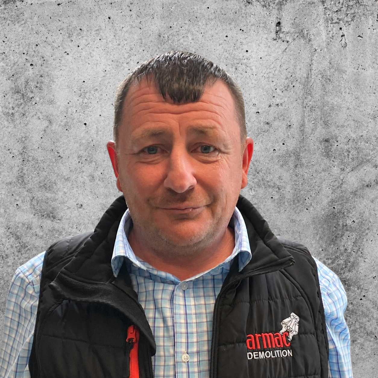 A middle-aged man in a checked shirt and black vest standing against a plain background.