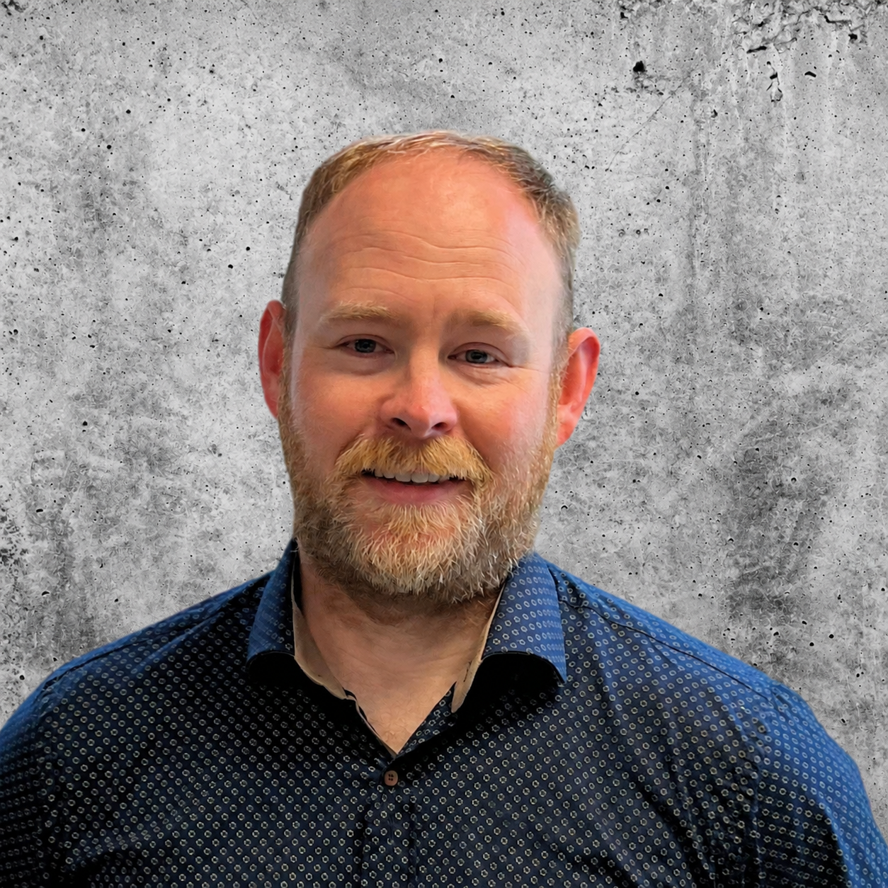 A man with a beard and short hair smiling, wearing a dark blue patterned shirt, standing against a plain gray background.