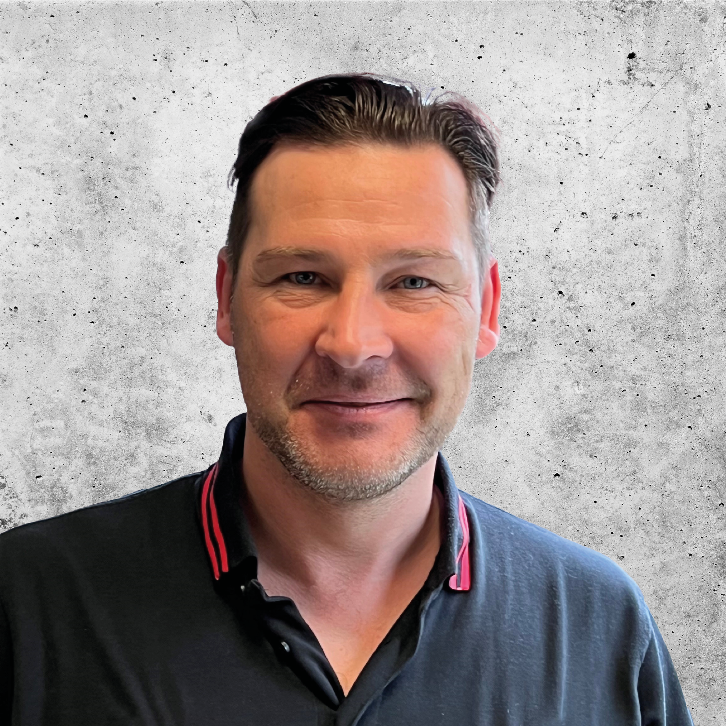 A smiling man with short dark hair and a beard, wearing a black polo shirt with red and white accents, standing against a plain grey background.