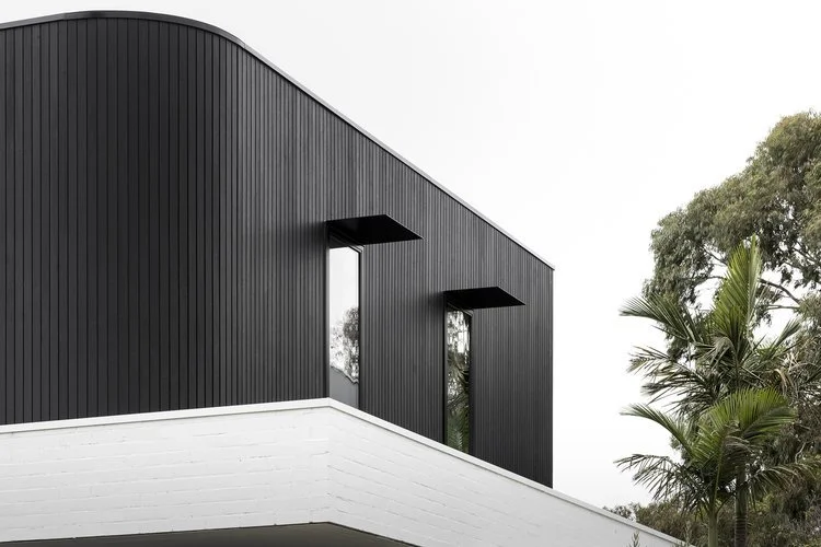 Modern architectural design featuring a black, vertically-paneled facade with large windows, against a white brick base and trees in the background.