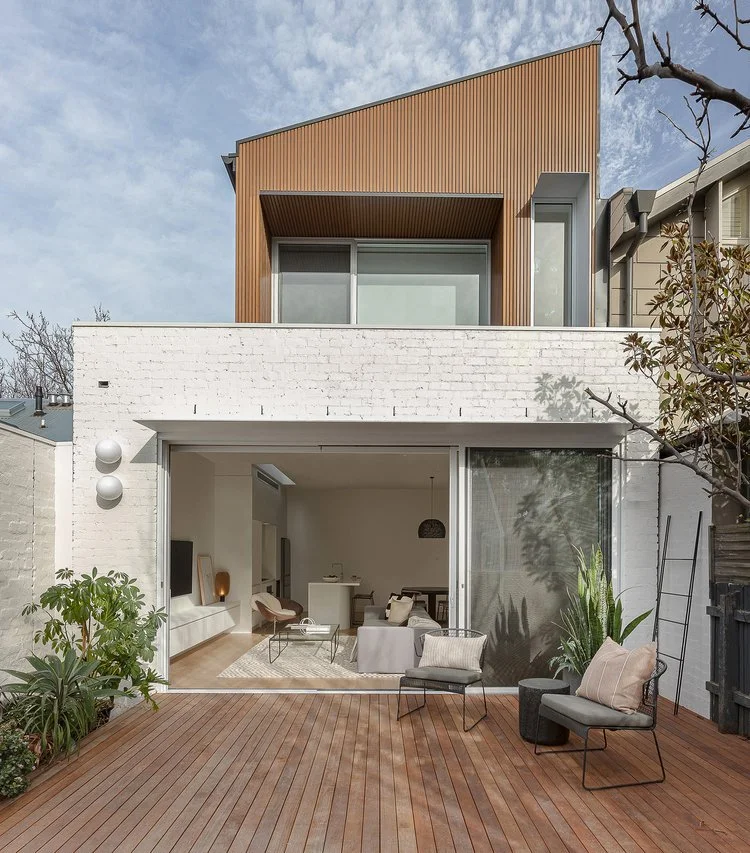 Modern house with white brick walls and timber accents, featuring a spacious outdoor deck furnished with chairs and a table, overlooking an interior living space visible through large glass sliding doors.
