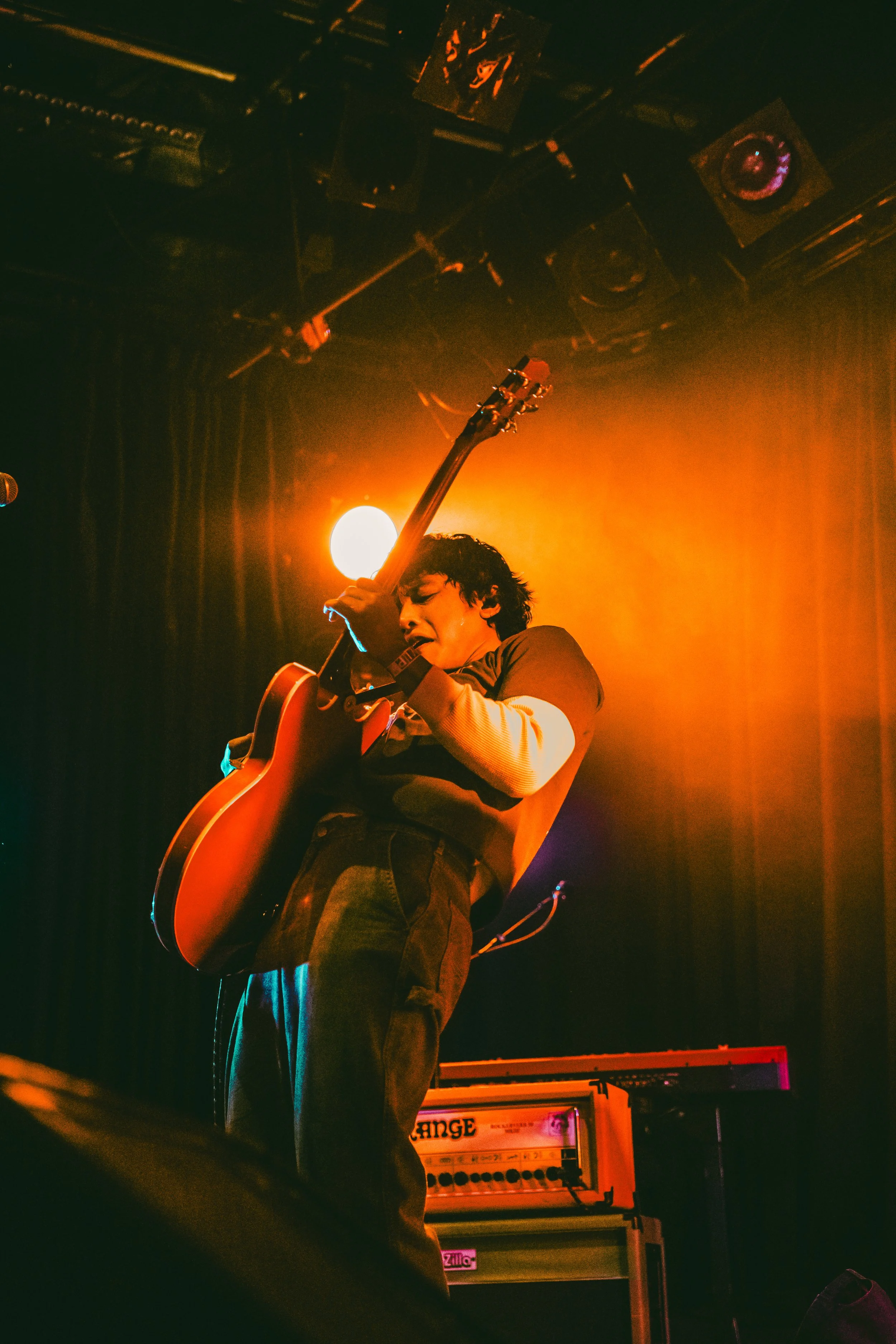 Musician performing on stage with an electric guitar, illuminated by warm stage lighting, and amplifiers visible in the background.