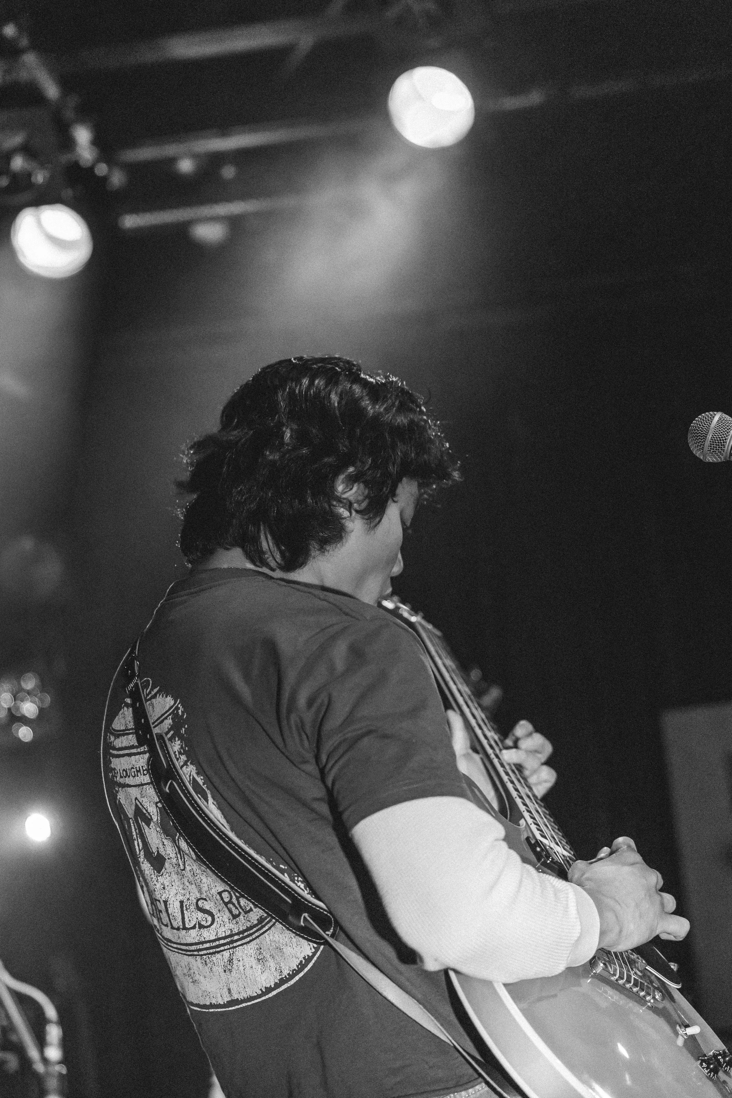 A person plays an acoustic guitar on stage, with stage lighting above and a microphone nearby, captured in black and white.