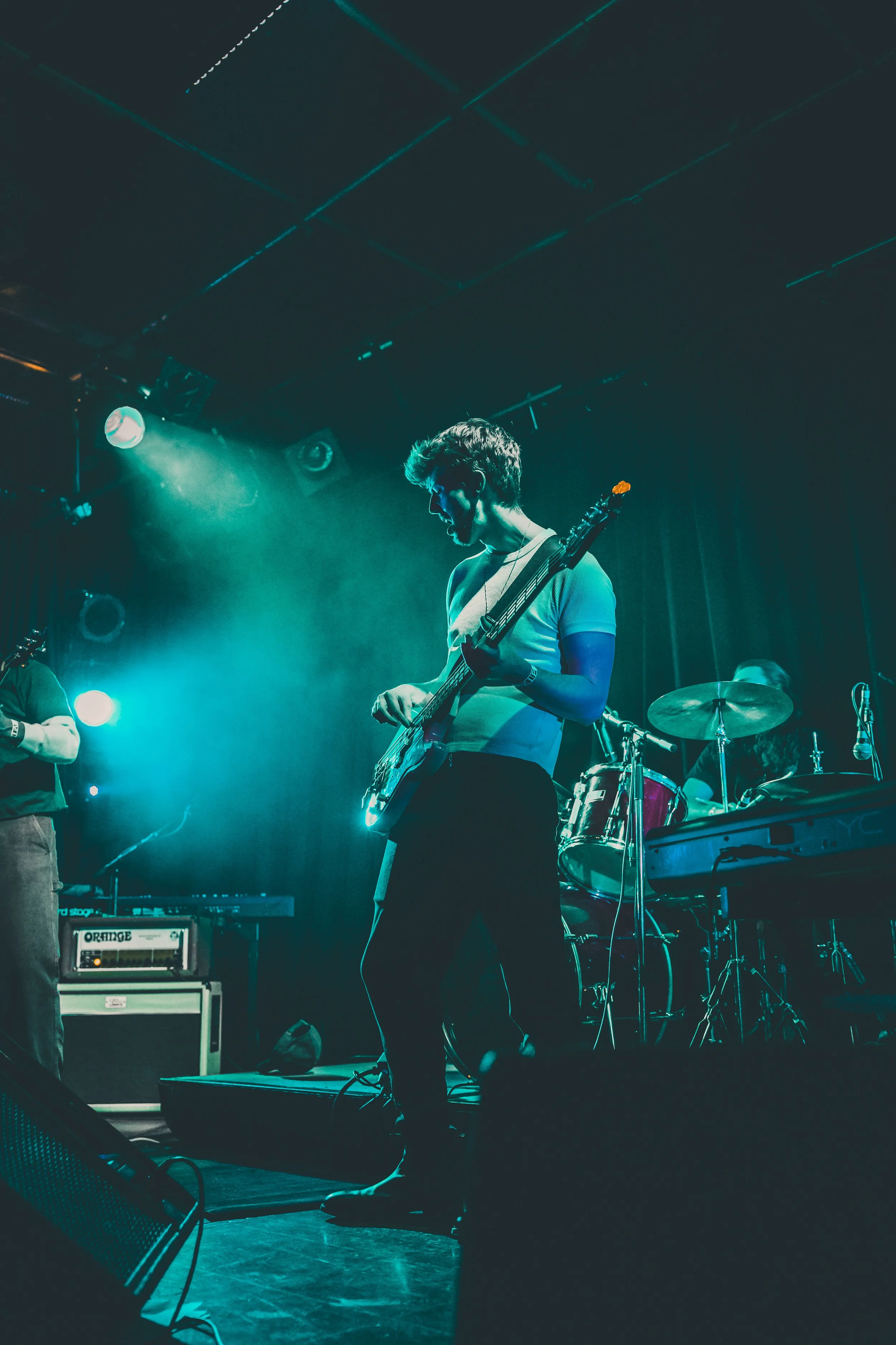 Young musician playing an electric guitar on stage under blue stage lighting with drums and keyboard in the background.