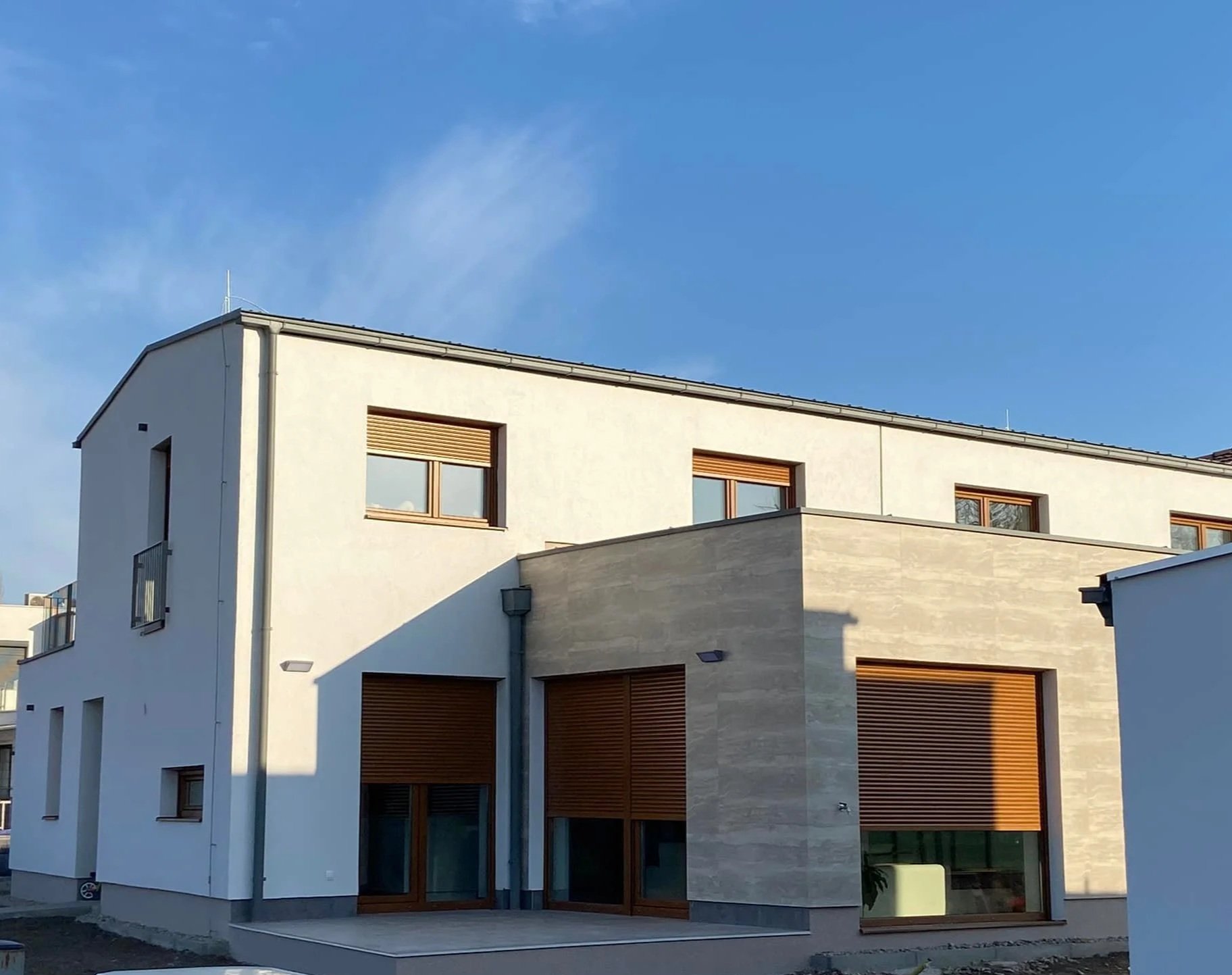 Modern two-story house with white and beige exterior walls, large windows with wooden shutters, and a flat roof, under a clear blue sky.