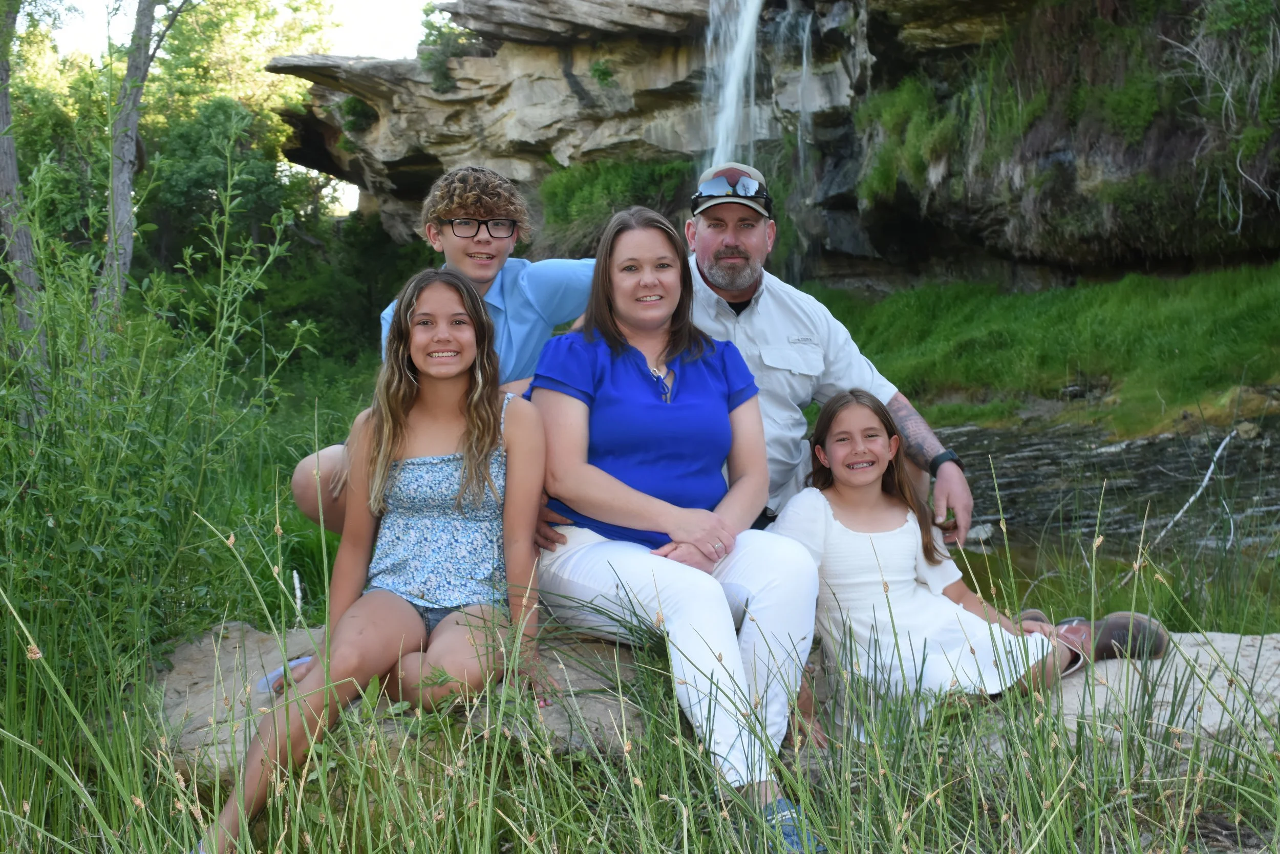 Family of six posing outdoors near a waterfall with green foliage and rocks.