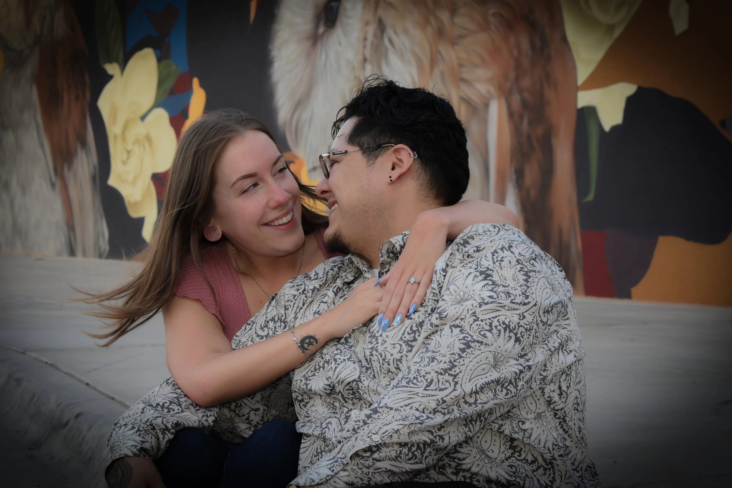A couple smiling and embracing each other outdoors, with a colorful mural of animals in the background.