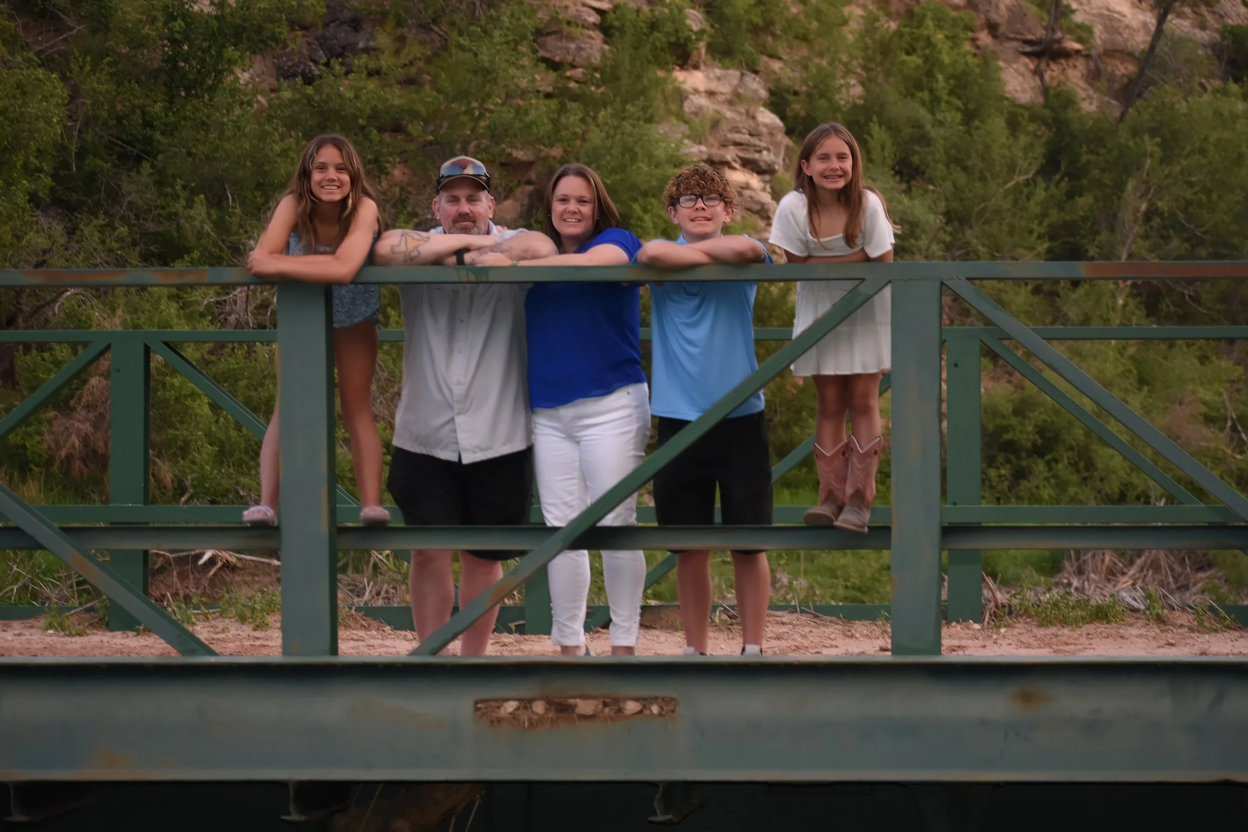 A family posing on a green metal bridge in a natural outdoor setting with trees and rocks in the background.