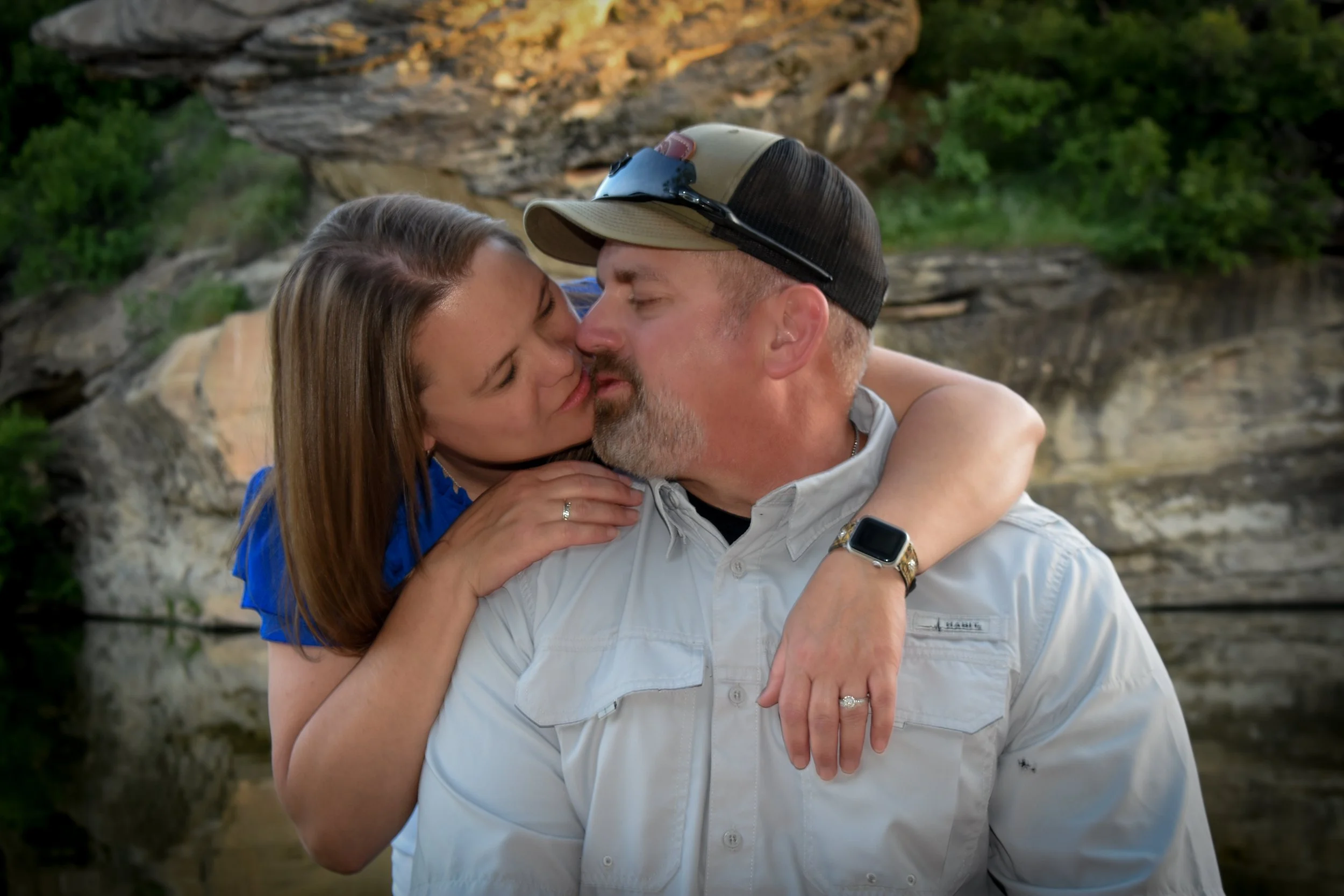 A couple sharing a kiss outdoors near a rocky water feature, with green foliage in the background.