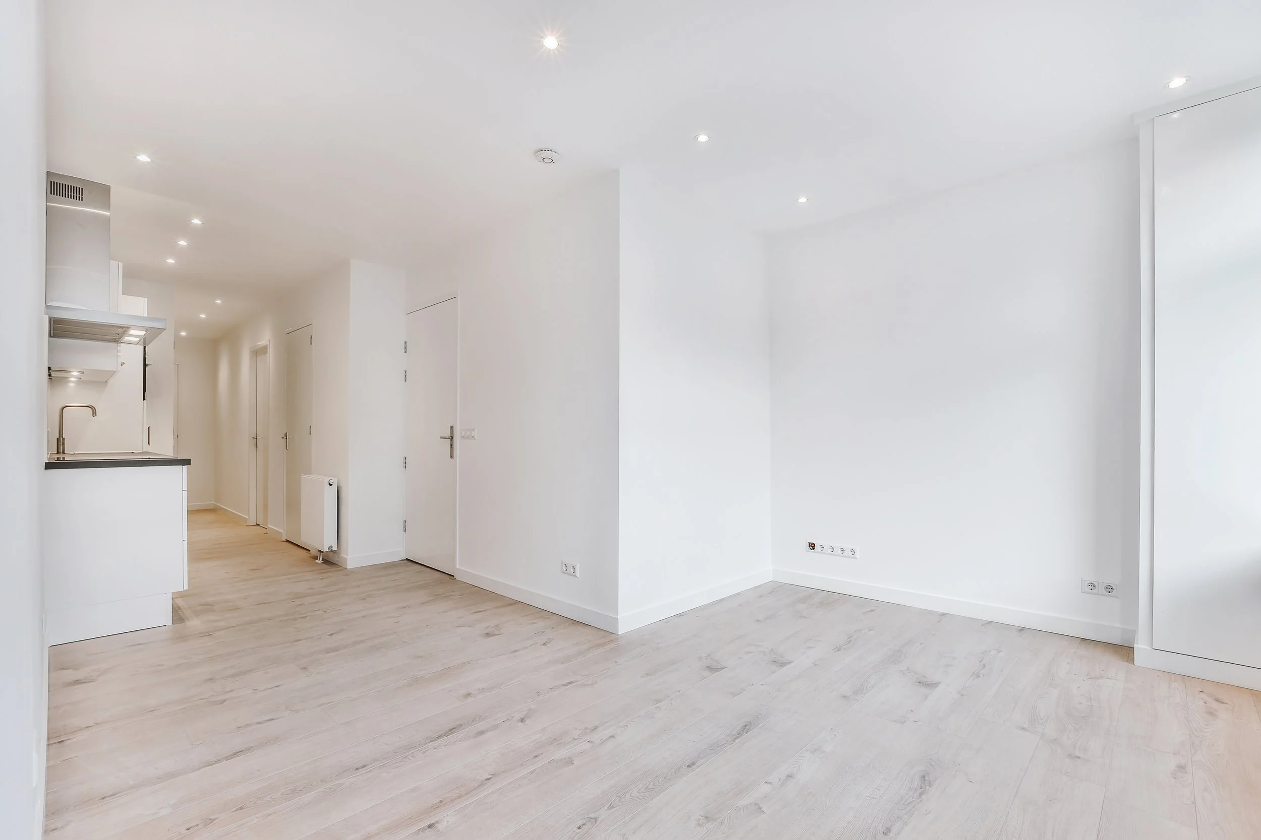 Ein leerer, heller Raum mit weißen Wänden, Holzboden, eingebauter Küche im Hintergrund und mehreren Deckenleuchten.