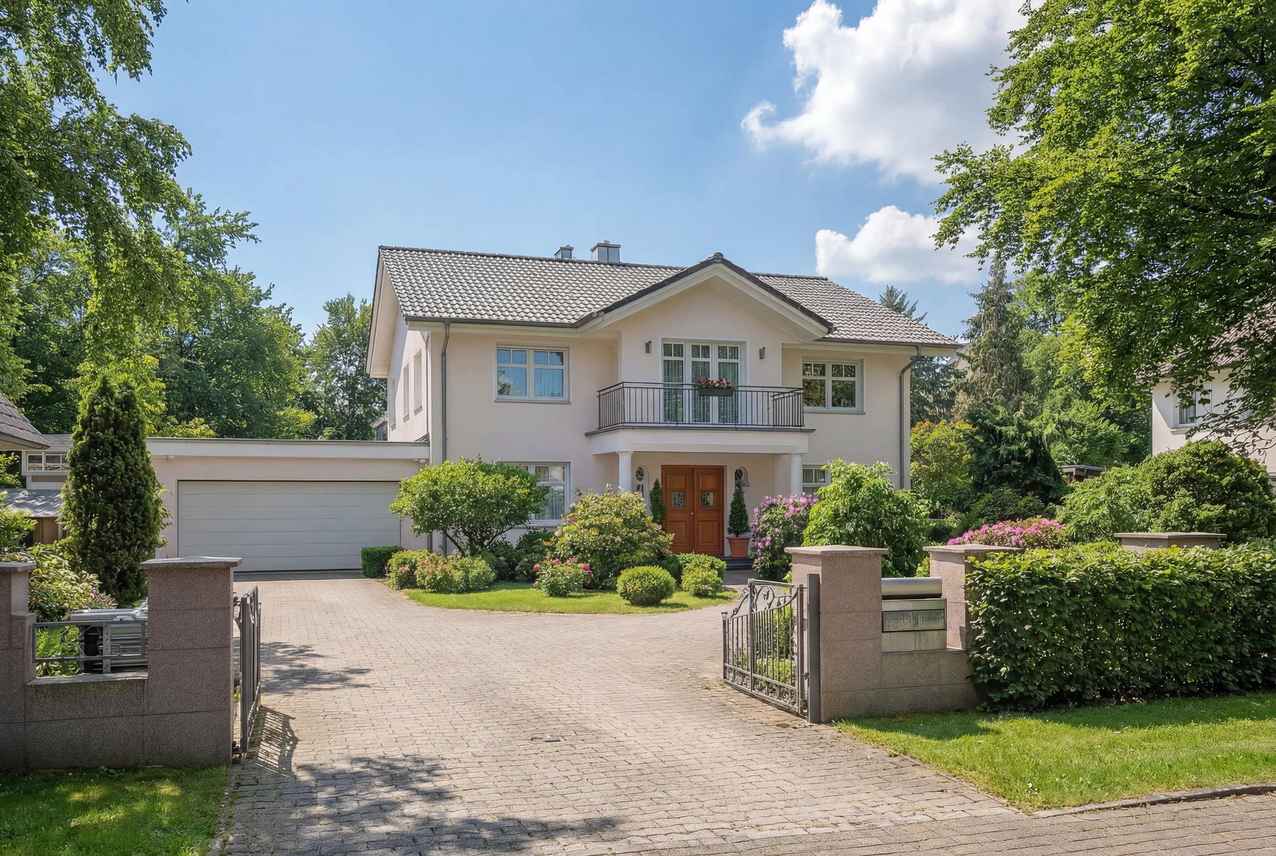 Ein zweistöckiges, weißes Haus mit Gitterfenstern, einer Holzhaustür und einem Balkon mit Blumenkasten, umgeben von einem Garten mit Sträuchern, Bäumen und Blumen, bei sonnigem Himmel.