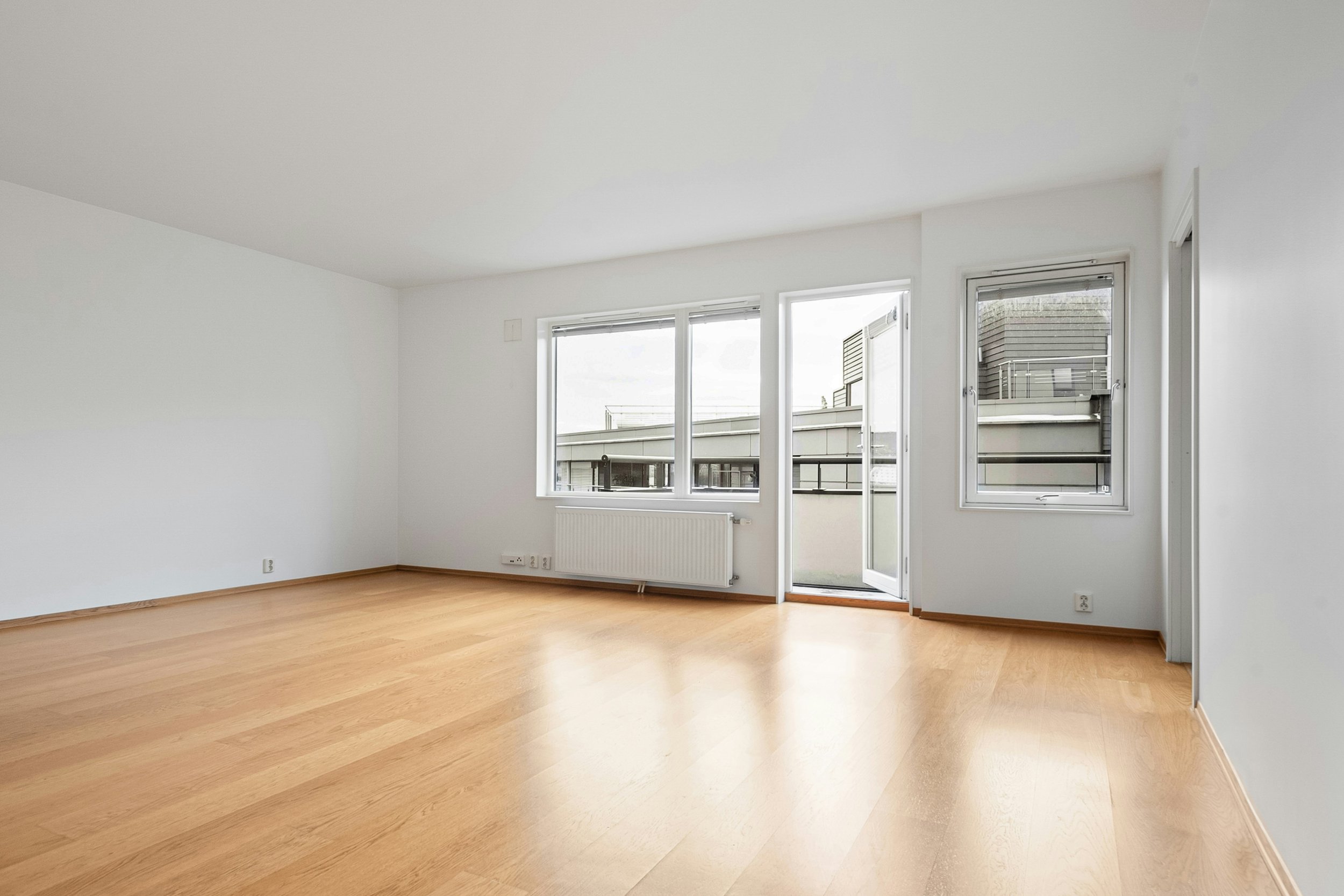 Leerer, heller Raum mit weißen Wänden, Holzboden, großen Fenstern und einer Tür, die auf einen Balkon führt.