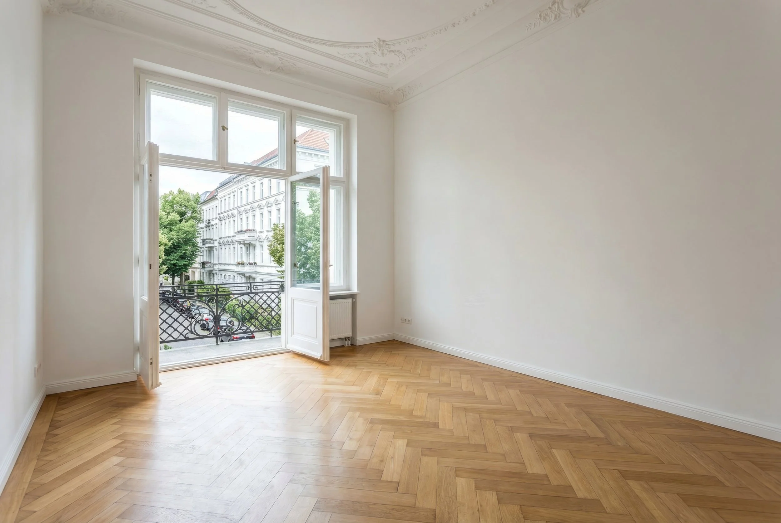 Ein leeres Zimmer mit weißen Wänden, Holzboden im Fischgrätmuster, großer Fensteröffnung mit Balkon und Blick auf eine historische Stadtstraße mit Bäumen und mehrstöckigen Gebäuden.
