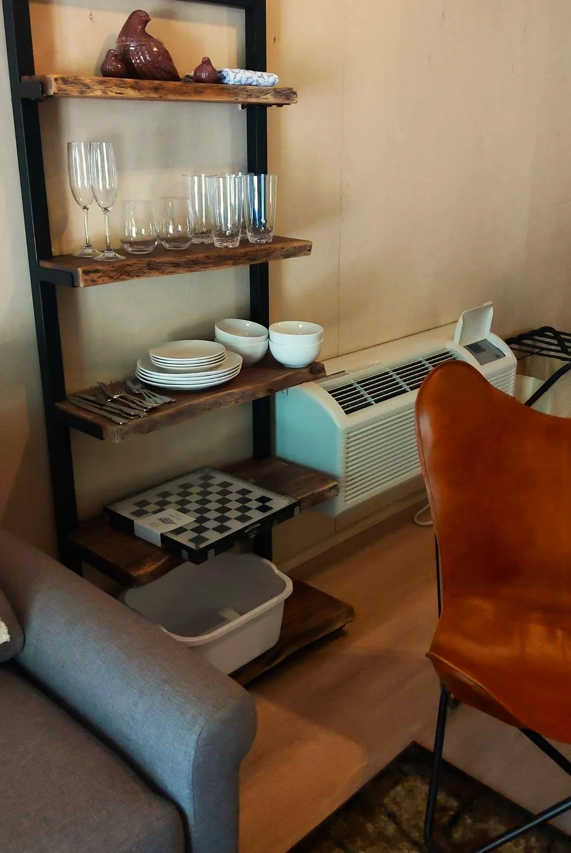Wooden shelving unit with glassware, plates, bowls, cutlery, and ceramic rooster, next to an air conditioner and a chair.