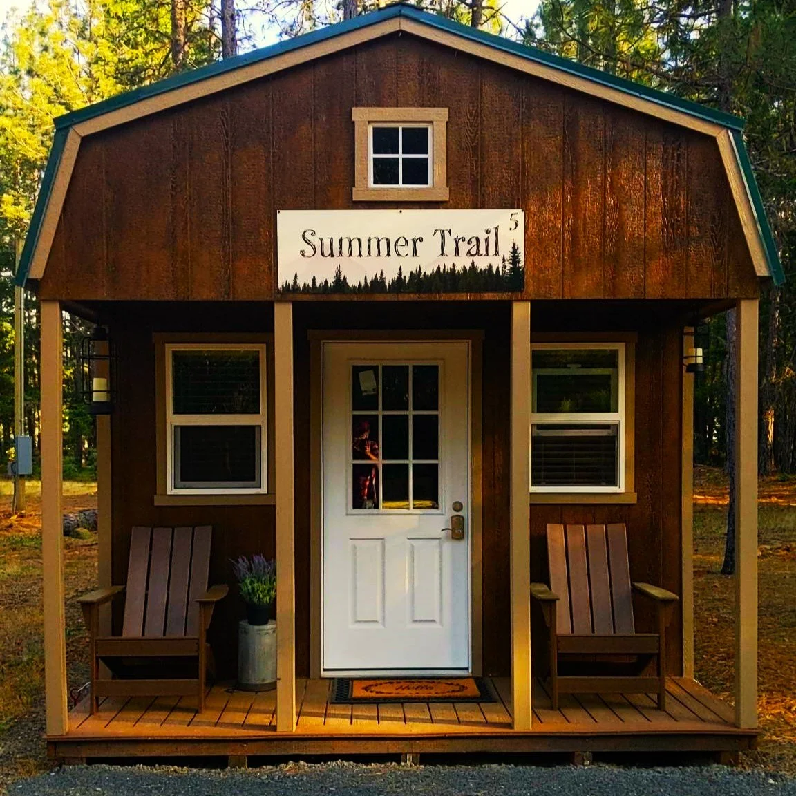 A small brown cabin with a white door and windows, featuring a porch with two wooden chairs. A sign above the entrance reads 'Summer Trail 5,' and the cabin is set in a wooded area.