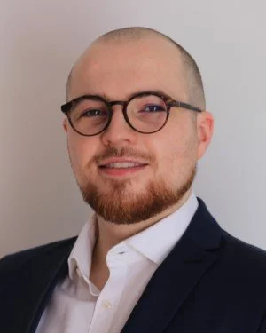 Porträt eines Mannes mit Brille, kurzer Haar- und Barttracht, trägt einen dunklen Anzug und ein weißes Hemd.