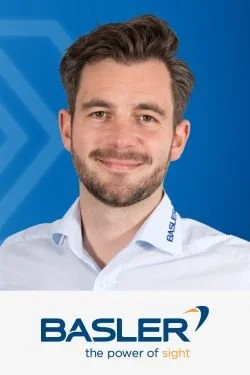 Porträt eines Mannes mit dunklem Haar und Bart, trägt ein weißes Hemd mit Basler Logo, im Hintergrund blaue Wand mit Basler Logo.