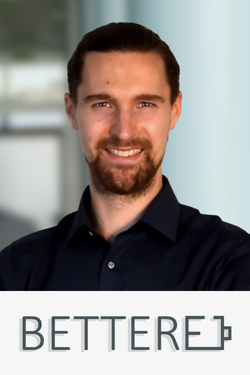 Ein Mann mit dunklem Haar, Bart und dunklem Hemd, der vor einem Büro hintergrund steht. Das Bild enthält den Text "BETTERE".