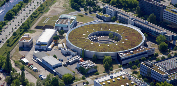 Luftaufnahme eines modernen Gebäudekomplexes mit einem großen runden Gebäude in der Mitte, das einen Innenhof hat, umgeben von mehreren anderen Gebäuden und viel Grünflächen.