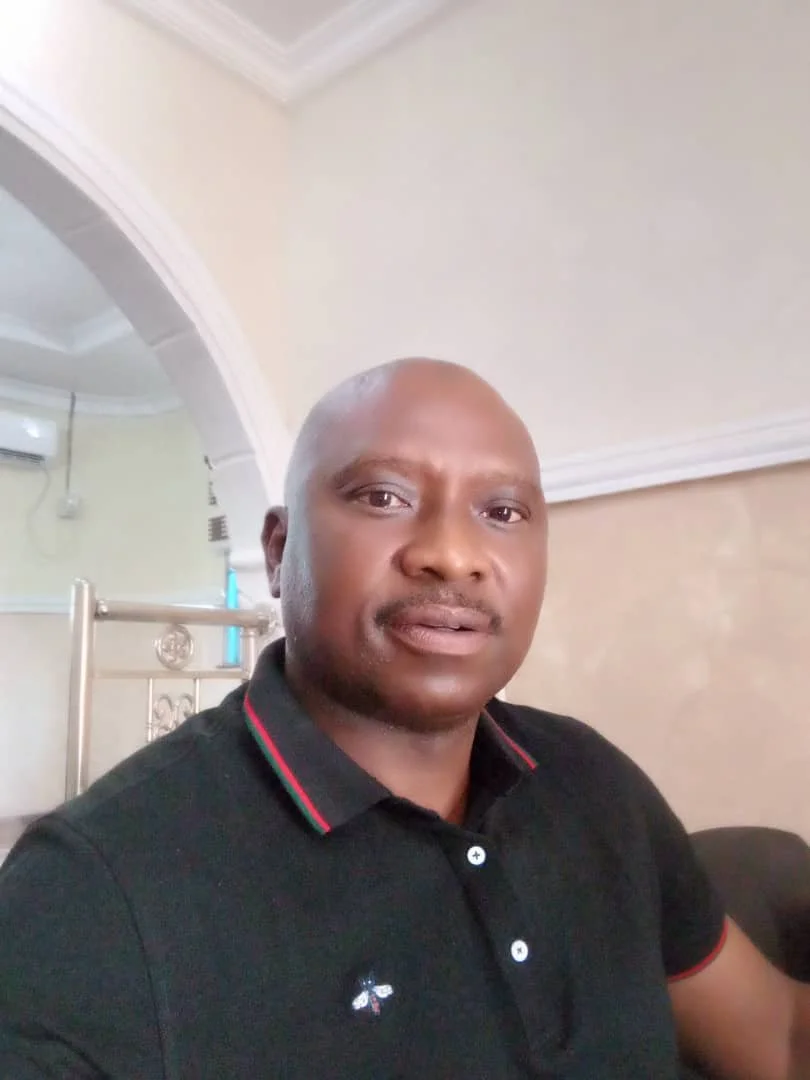 A man with a shaved head wearing a black polo shirt with red and green stripes on the collar, sitting indoors in front of a plain off-white wall and a partial view of a sofa.