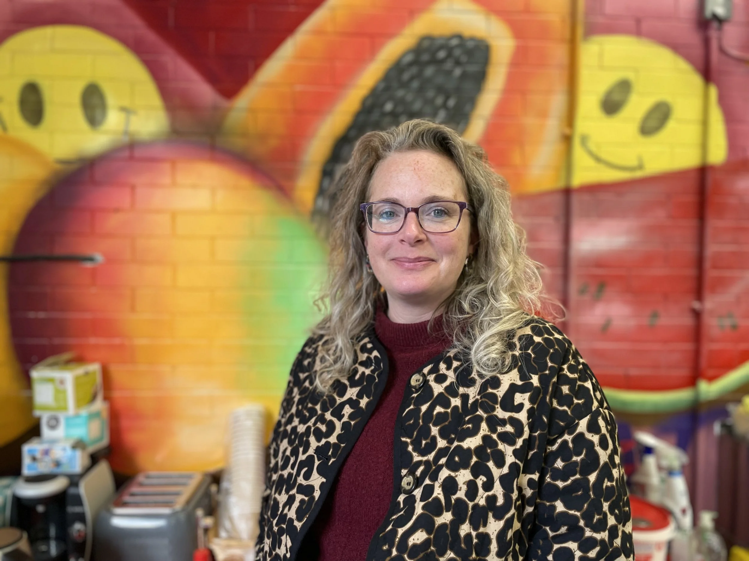 Photo of Davina looking at the camera and smiling, with colourful wall painted with fruit behind her