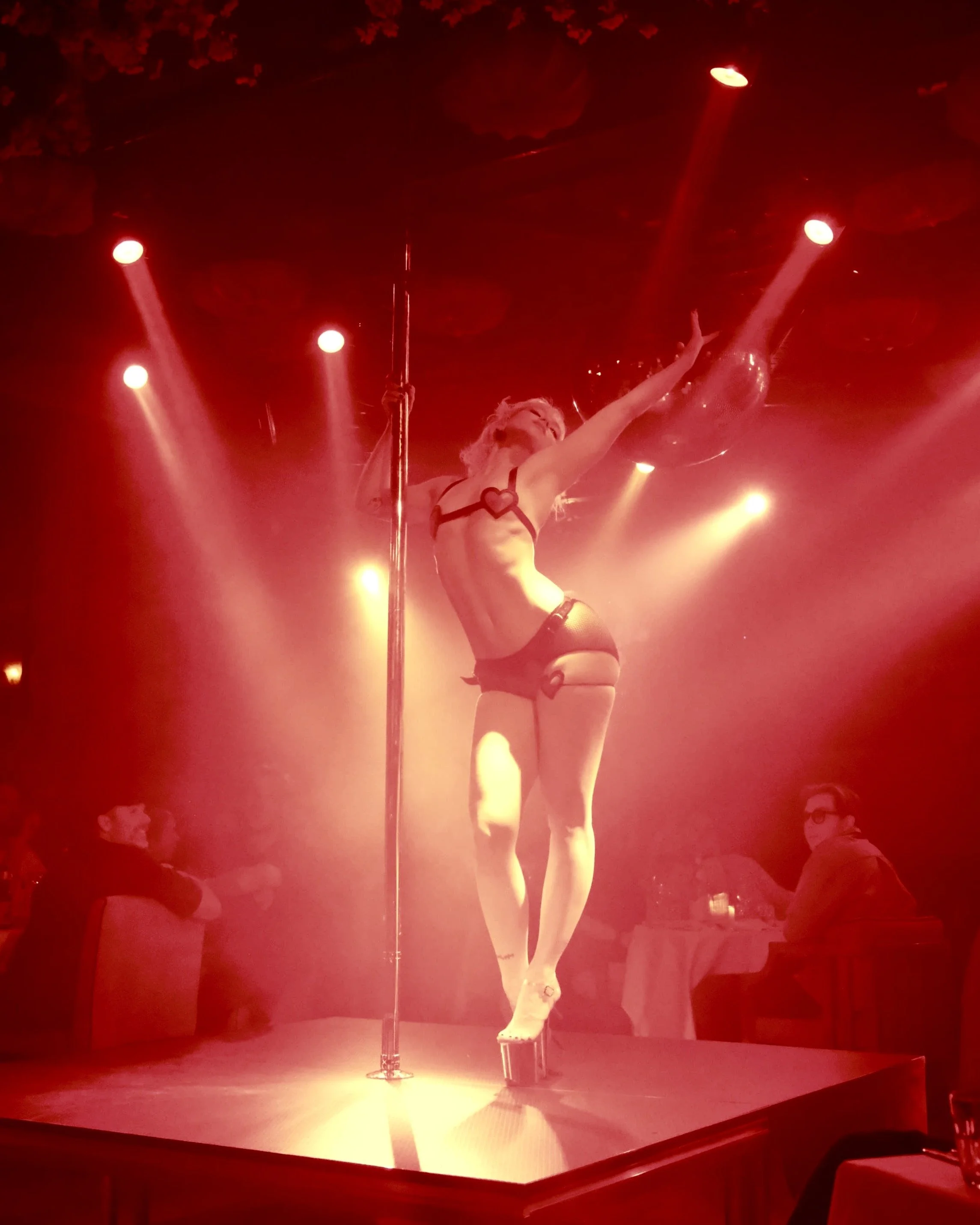 A woman performing a pole dance on stage under red lighting, wearing a black harness with a heart design, black shorts, and high heels. She has her arms raised and is balancing on the pole, with audience members visible in the background.
