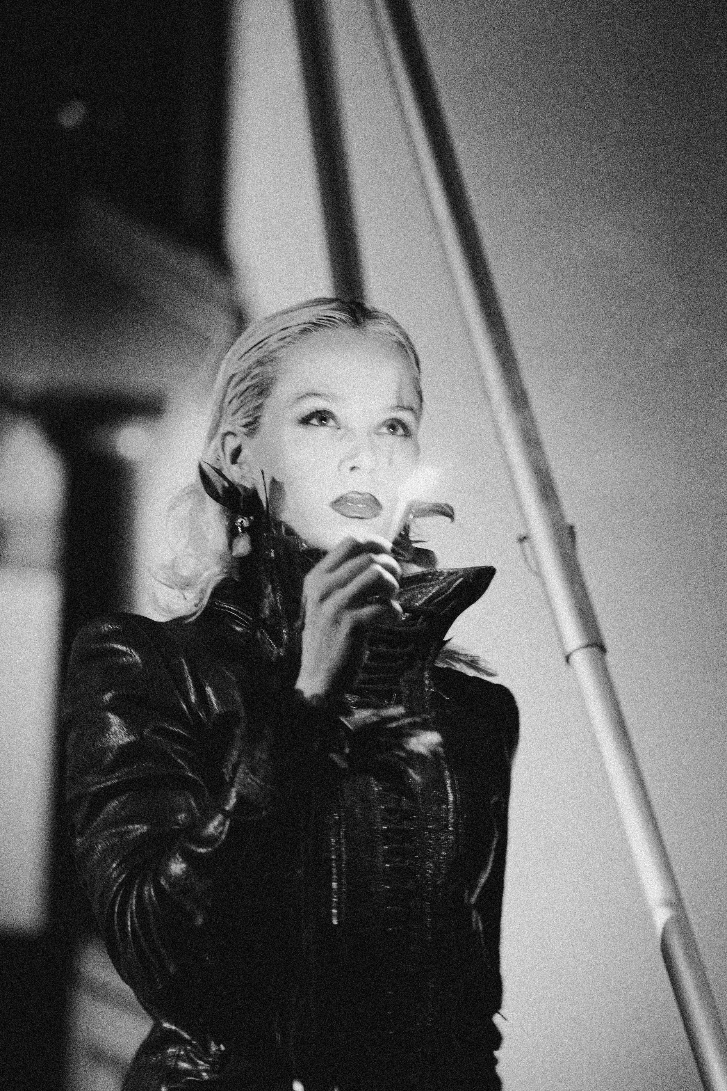 Black and white photo of a woman with platinum blonde hair, wearing a leather jacket and holding sunglasses, looking up with a confident expression.