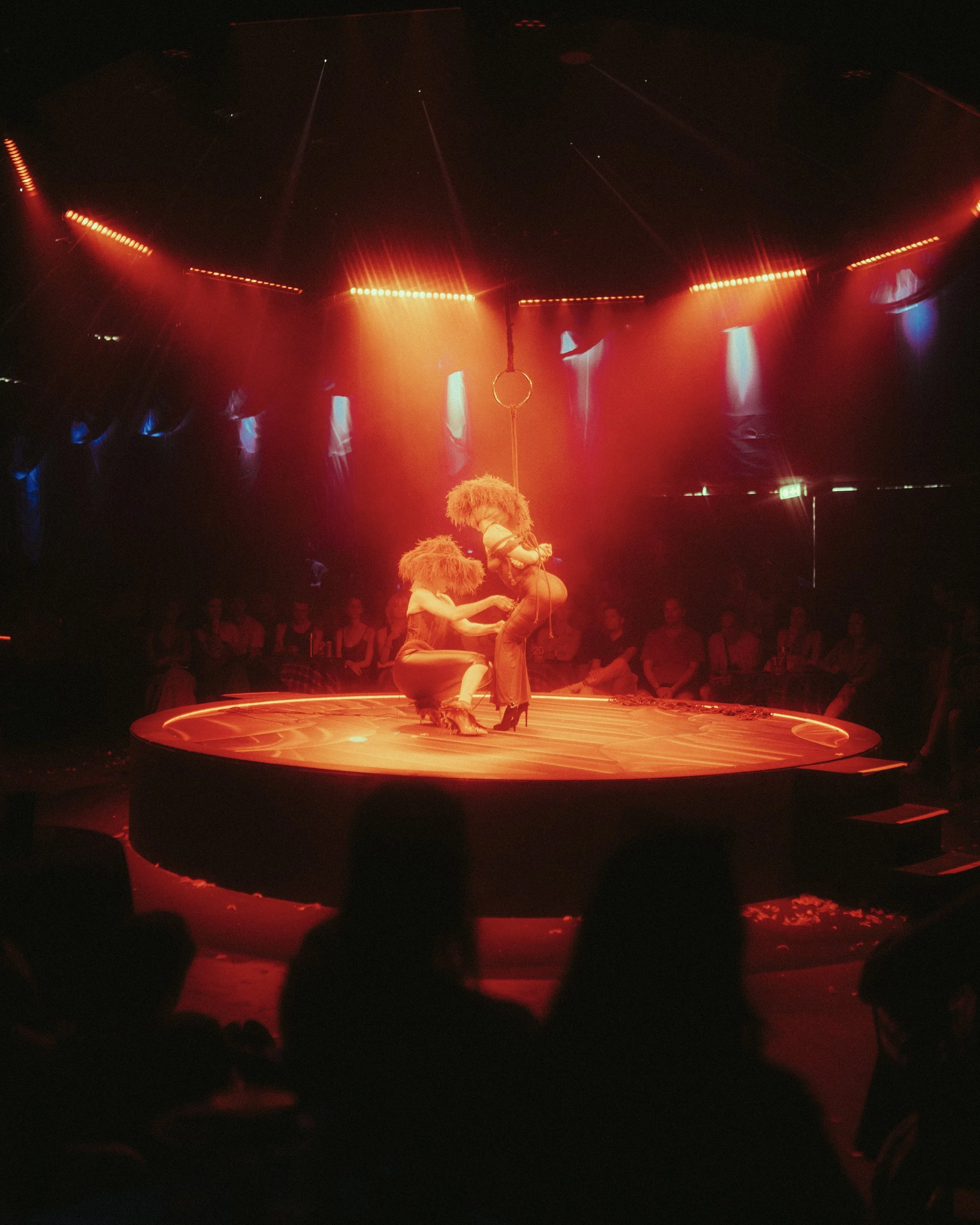 Acrobats performing a balancing act on a circus stage illuminated with red and blue lights, with an audience watching in the background.