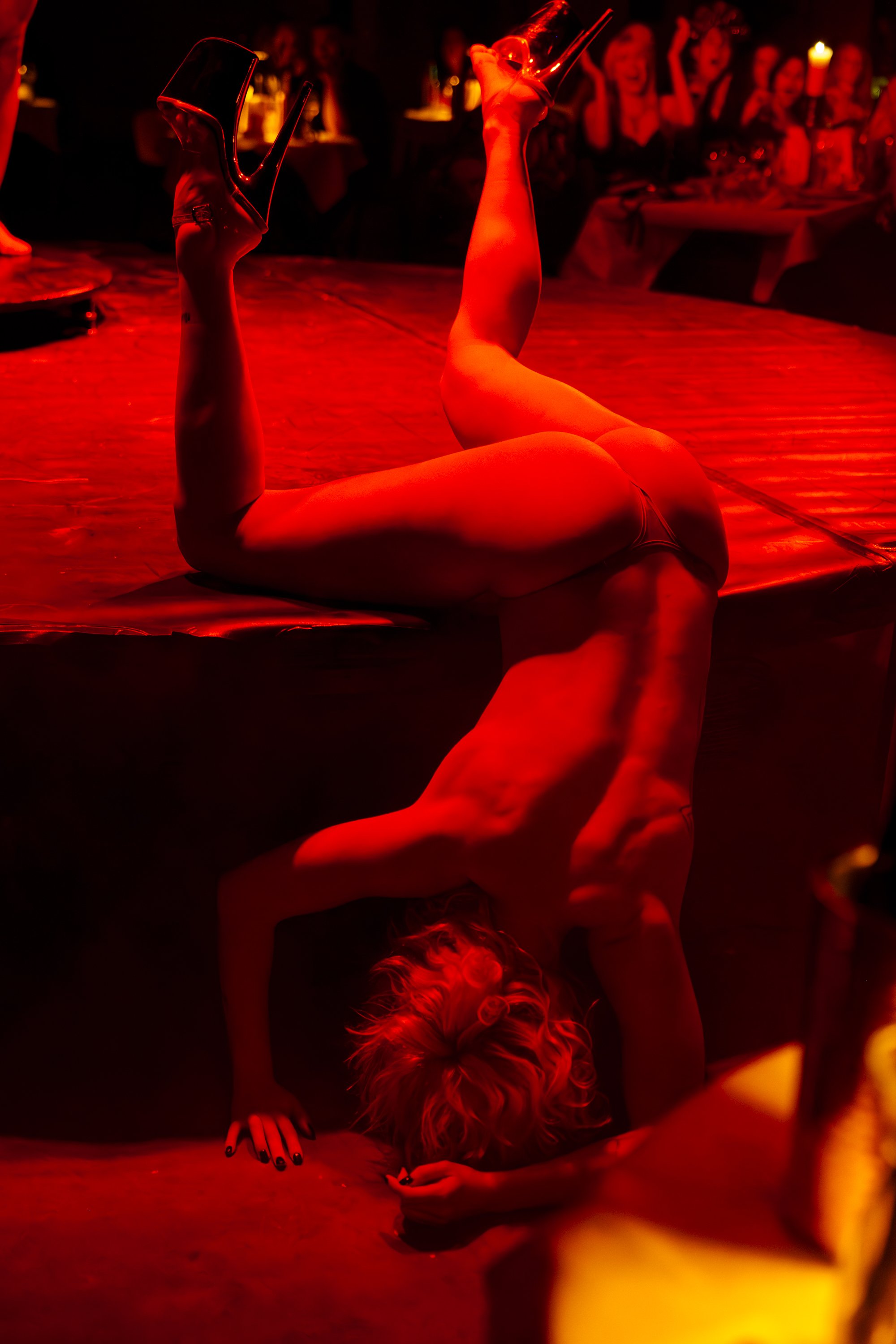 A person is performing a contortion pose on the floor with their hands and head touching the ground and wearing high heels, surrounded by an audience in a dimly lit, red-hued environment.