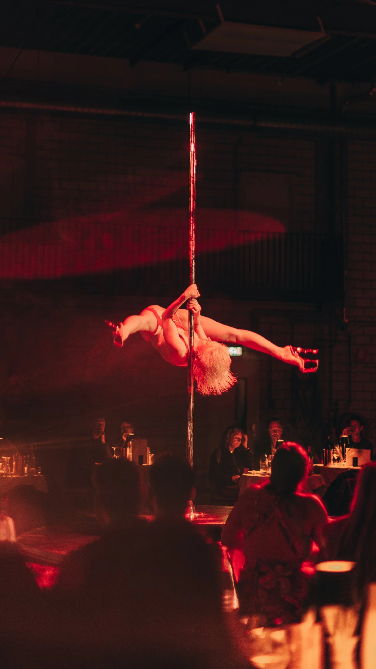 A female pole dance performer in Amsterdam on stage in a dimly lit venue with an audience watching performing an acrobatic trick.