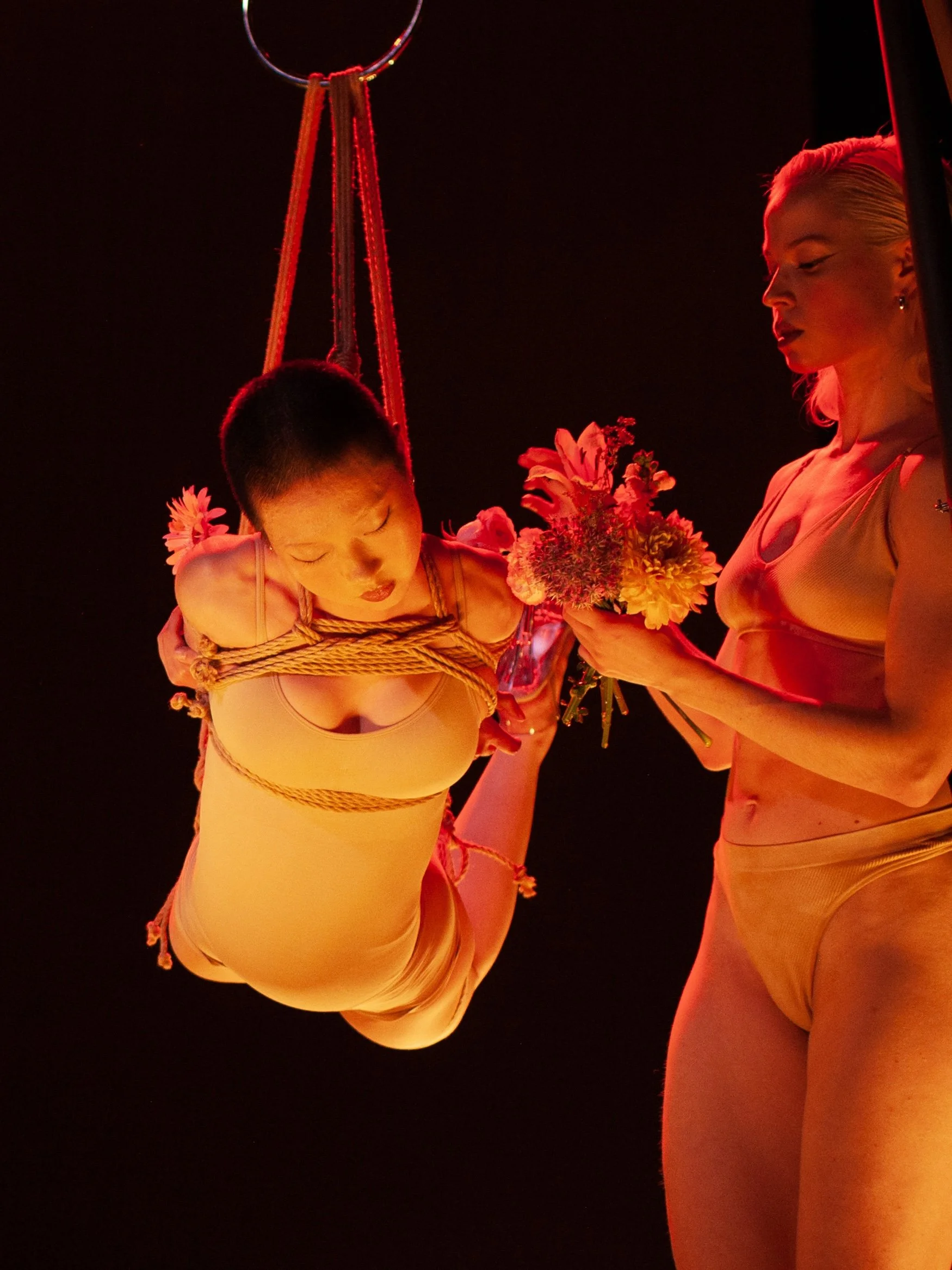 A person with light skin and blonde hair is holding a bouquet of flowers and is bound with ropes, with another person nearby holding the flowers. The scene is illuminated with red lighting.