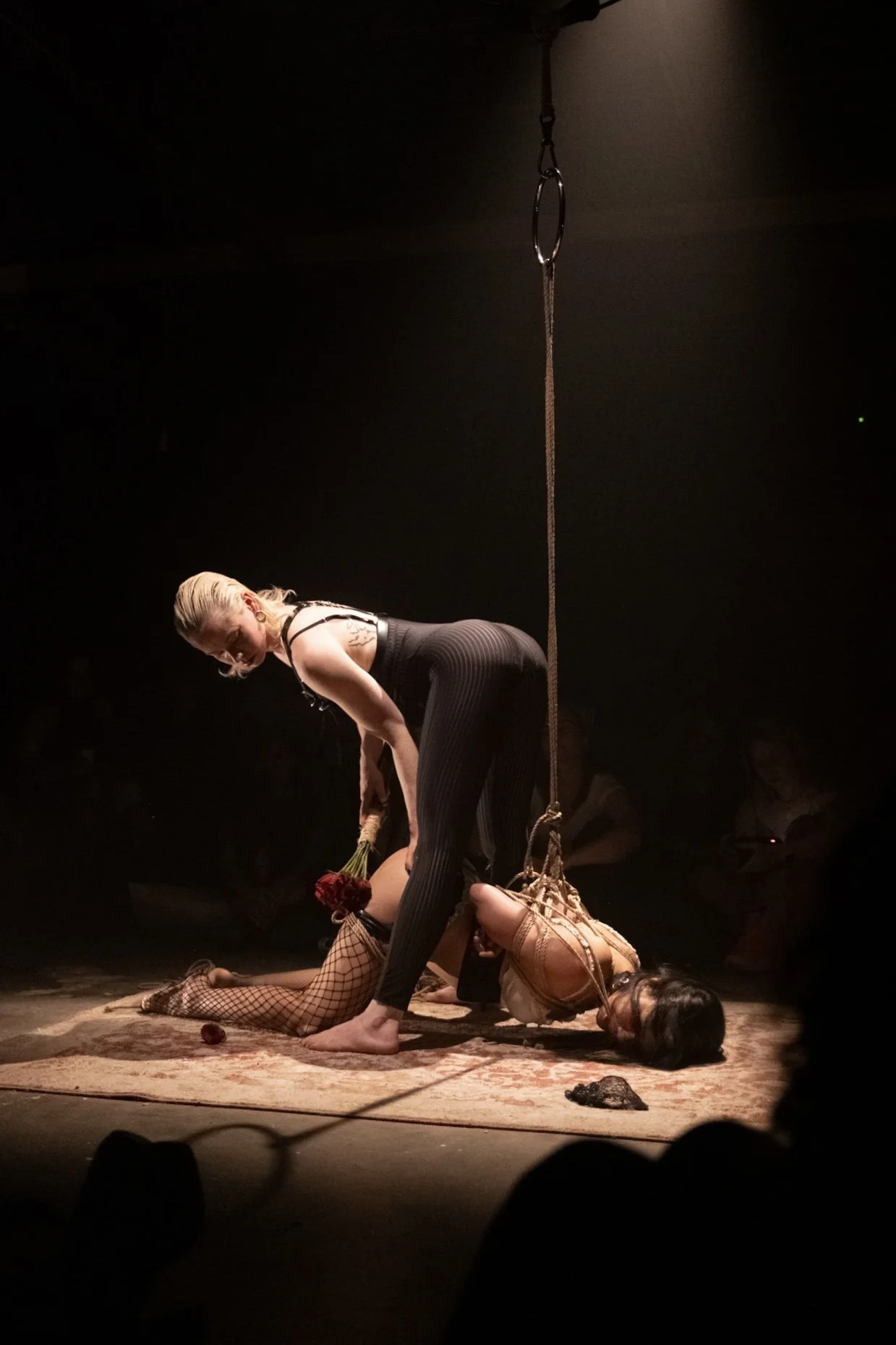 Two women performing a shibari japanese rope bondage performance on stage with dark background. One woman with blonde hair holds red roses and is bending forward, while the other woman in fishnet stockings is lying on the floor.