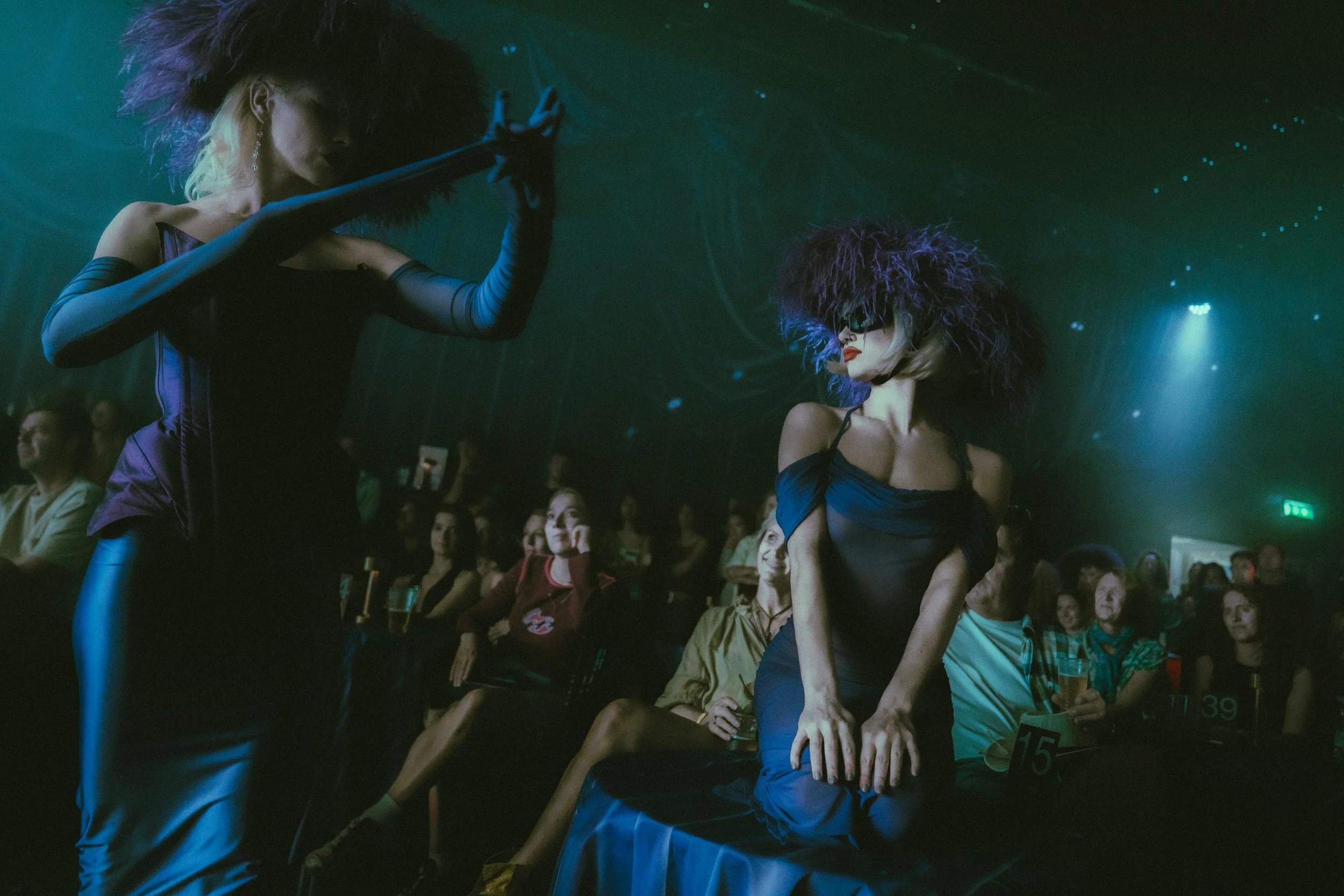 Two dancers with large, curly hats and dark outfits perform for an audience in a dimly lit nightclub with a starry background.