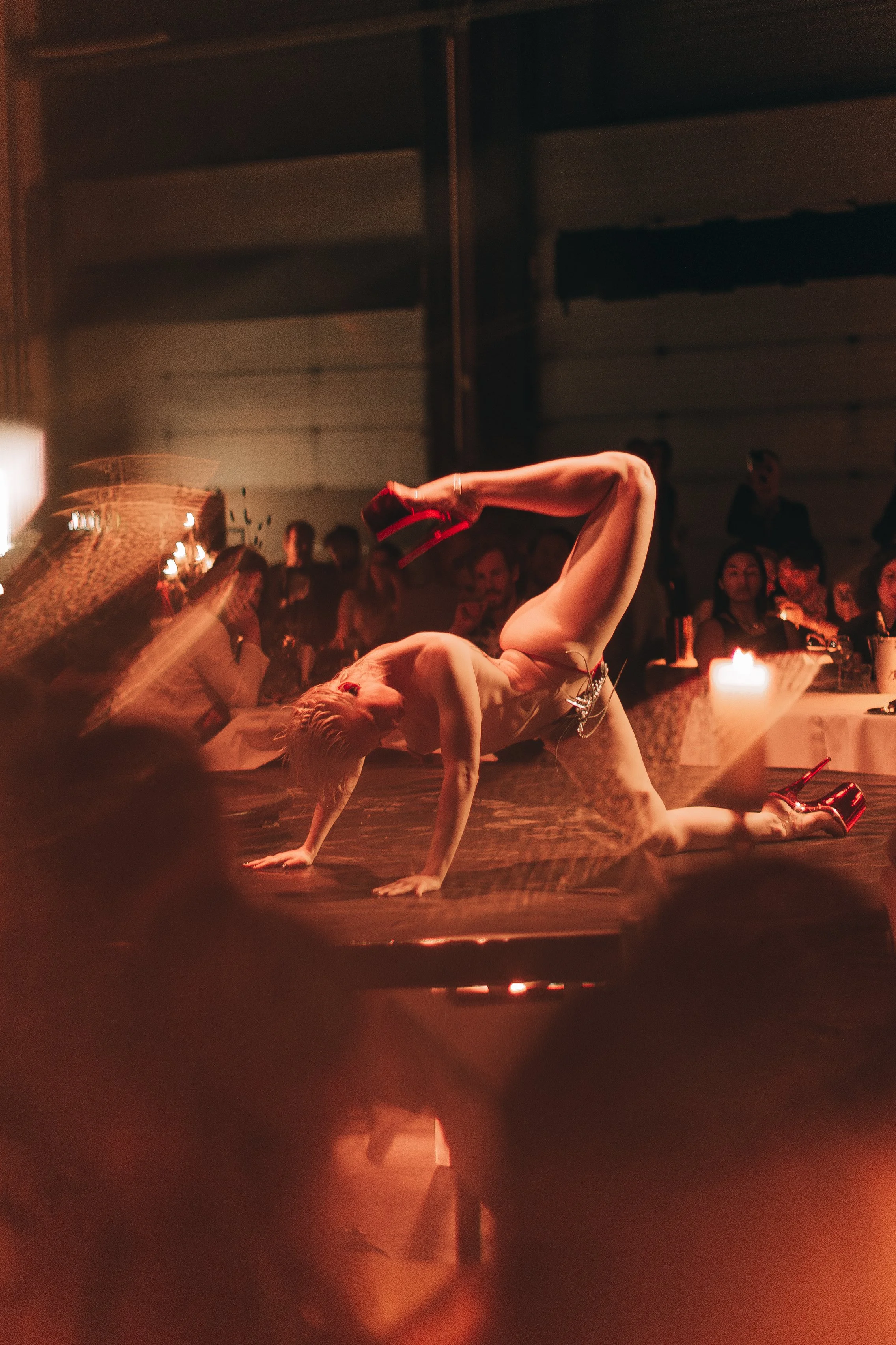 A performer in lingerie and high heels is on her hands and knees on a stage, engaging in a dance or acrobatic routine, with an audience seated at tables watching.