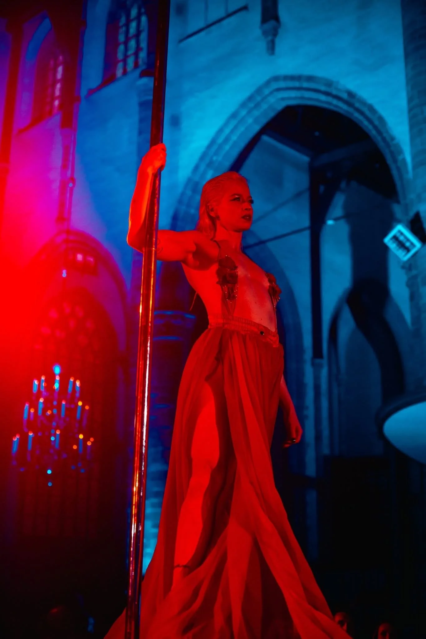 A woman with blonde hair is standing in front of a blue-lit brick wall, holding a vertical pole with her right hand. She wears a flowing orange dress with a sheer overlay and appears to be performing or posing.