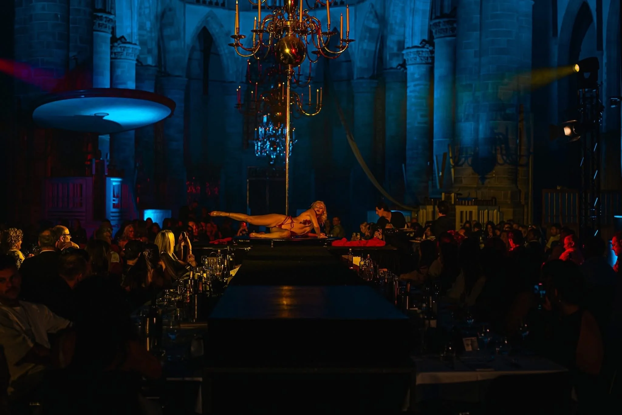 A performer dressed in revealing clothing lying on a stage in a grand hall with a large chandelier hanging from the ceiling, surrounded by an audience watching an entertainment show.