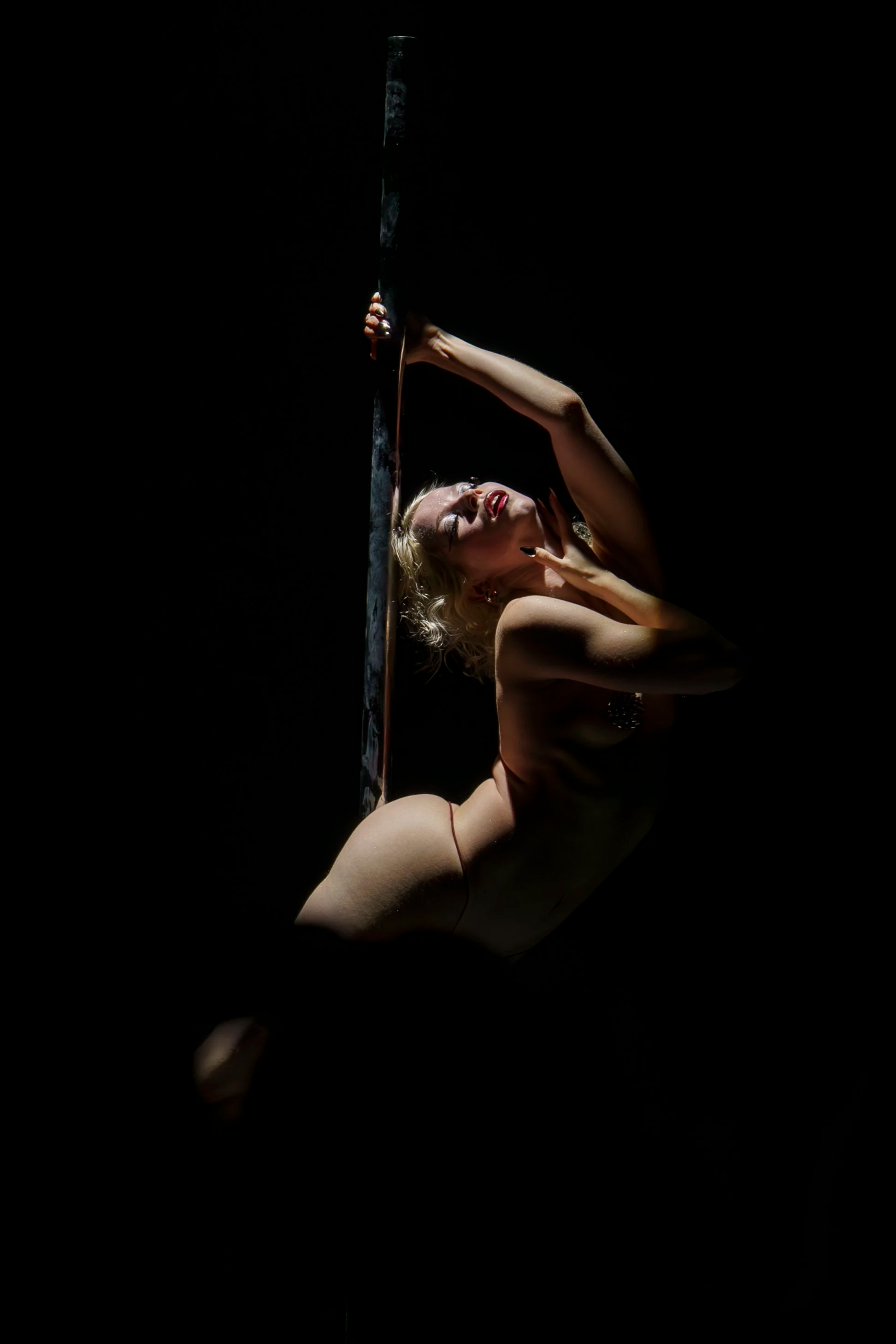 A woman with blond hair and makeup is pole dancing on stage against a dark background, holding a long vertical object, possibly a mirror or a sword.
