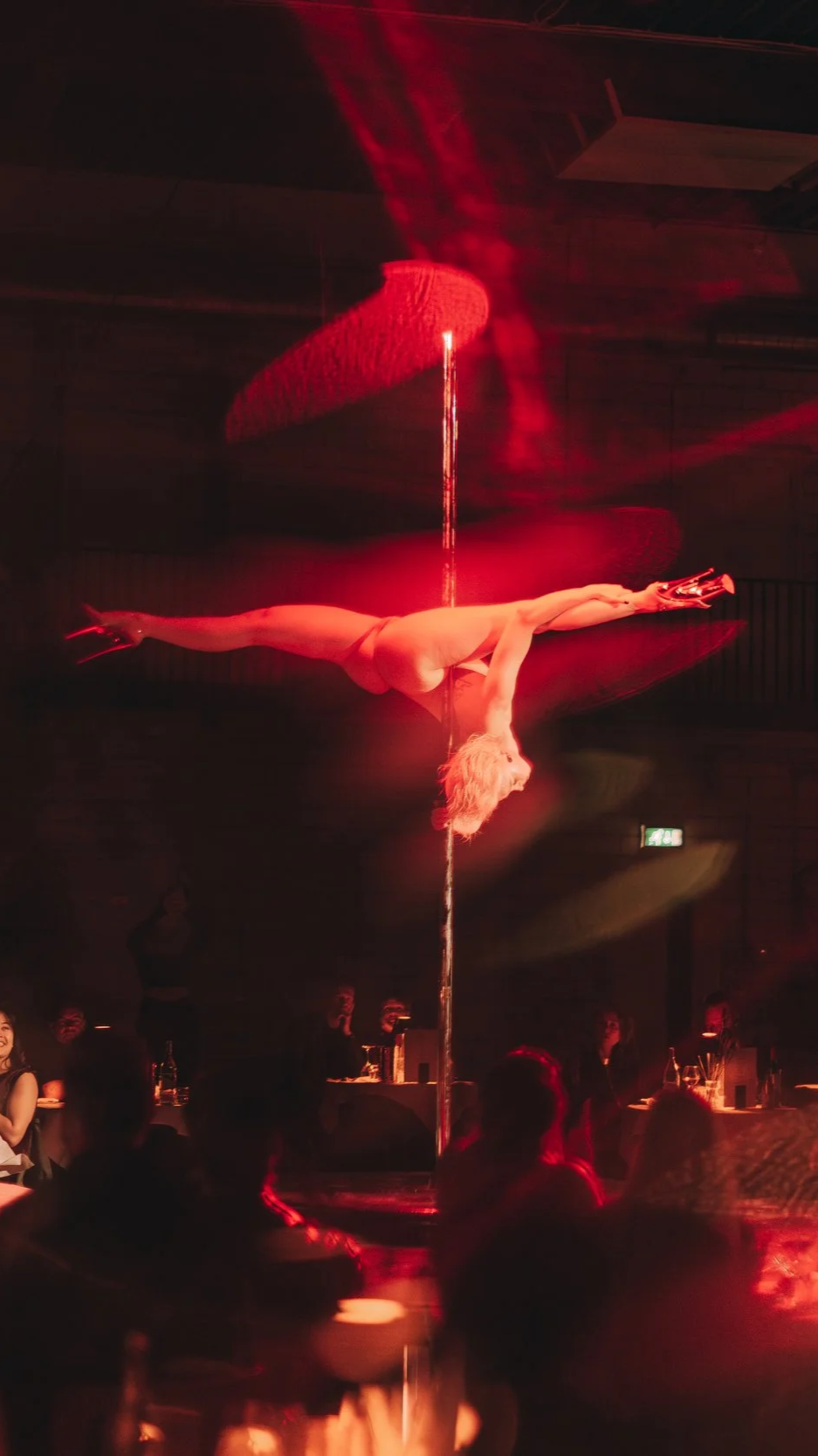 A female aerial performer in nude costume performs an acrobatic pose on a vertical pole during a show, illuminated by red stage lighting with audience members watching.