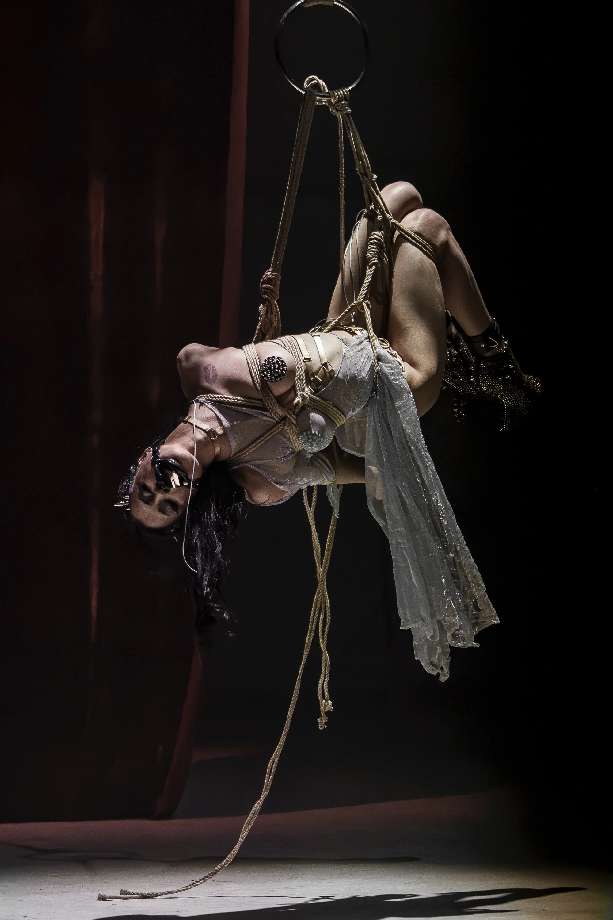 Shibari performance Amsterdam. Woman has been tied in a harness hanging from a suspension ring on stage.