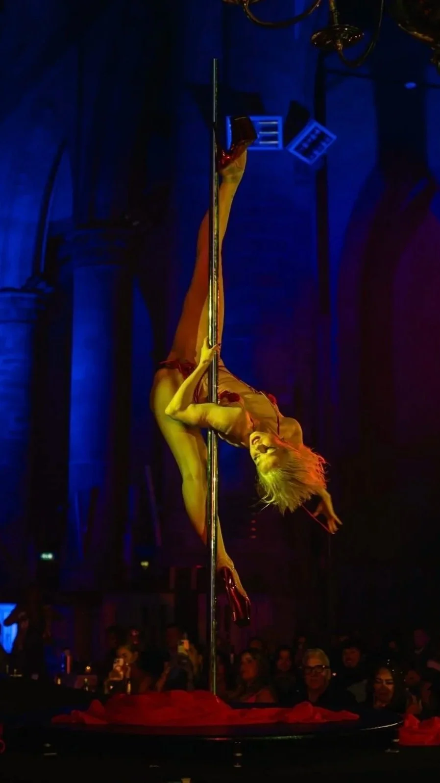 A female performer in a red dress and high heels performing a pole dance on stage in front of an audience.