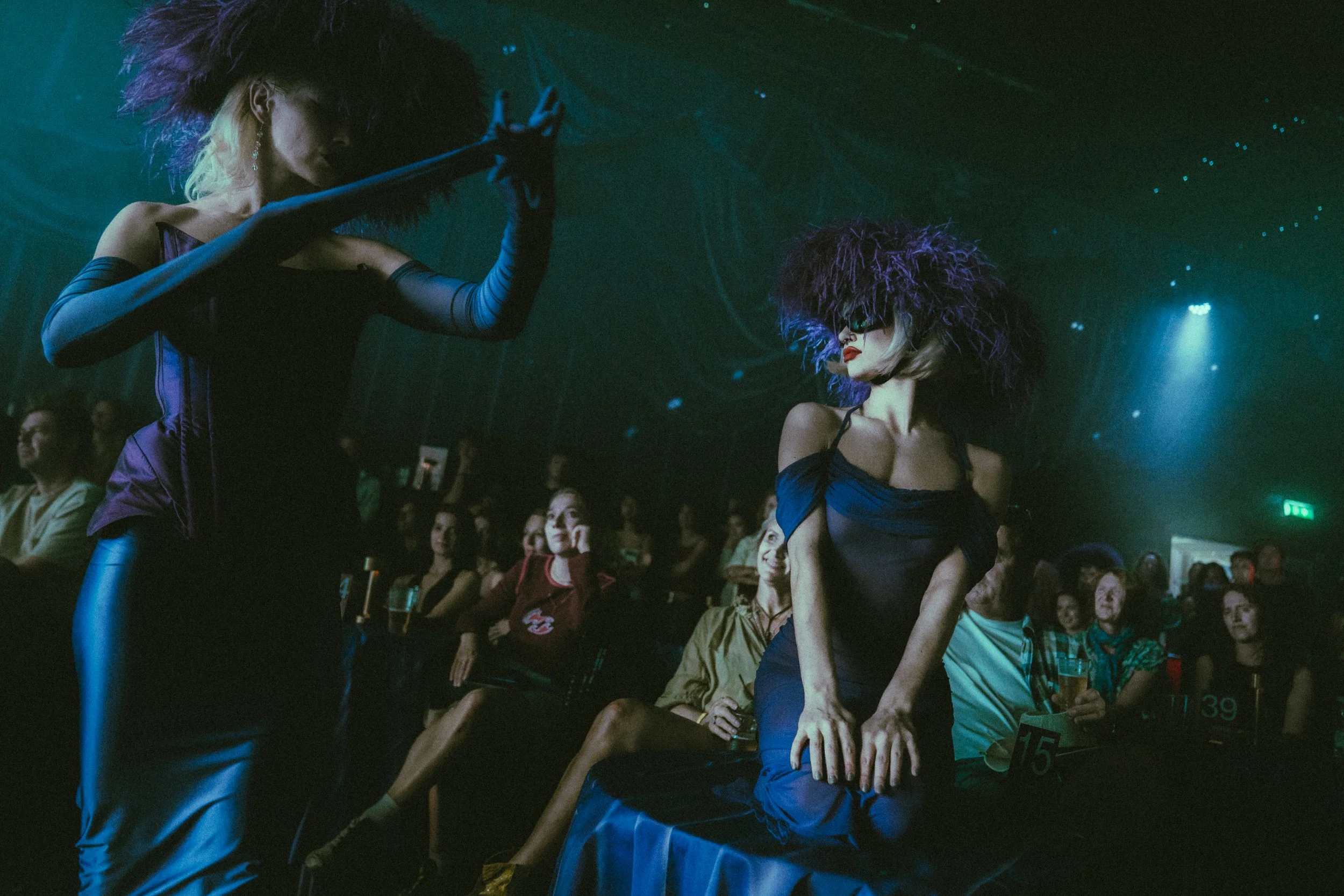Two women with big, curly wigs and fashionable outfits perform on stage in front of an audience at a nightclub. The audience is seated and watching the performance.