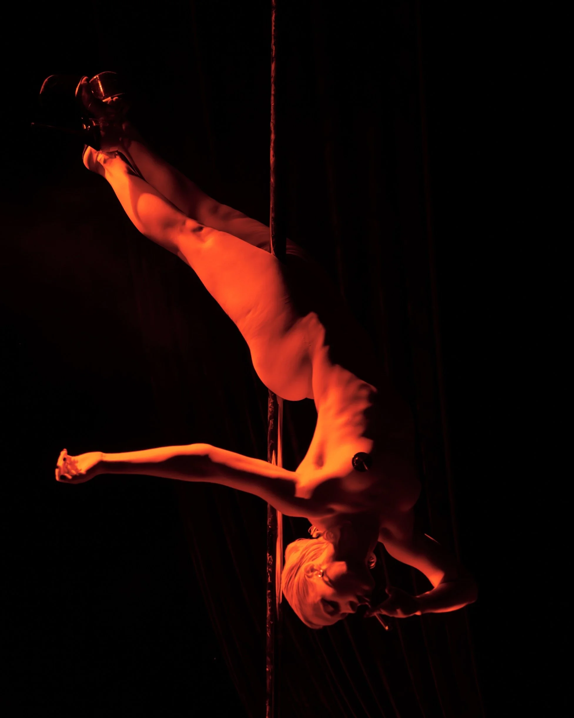 A person performing a pole dance move in a dark room, illuminated by red lighting, standing with one arm raised above their head and the other extended outward, with flowing fabric around them.