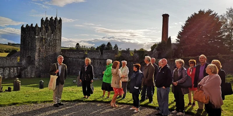 Group of people standing outdoors near a historic castle wall with a tower, some are talking, on a sunny day.