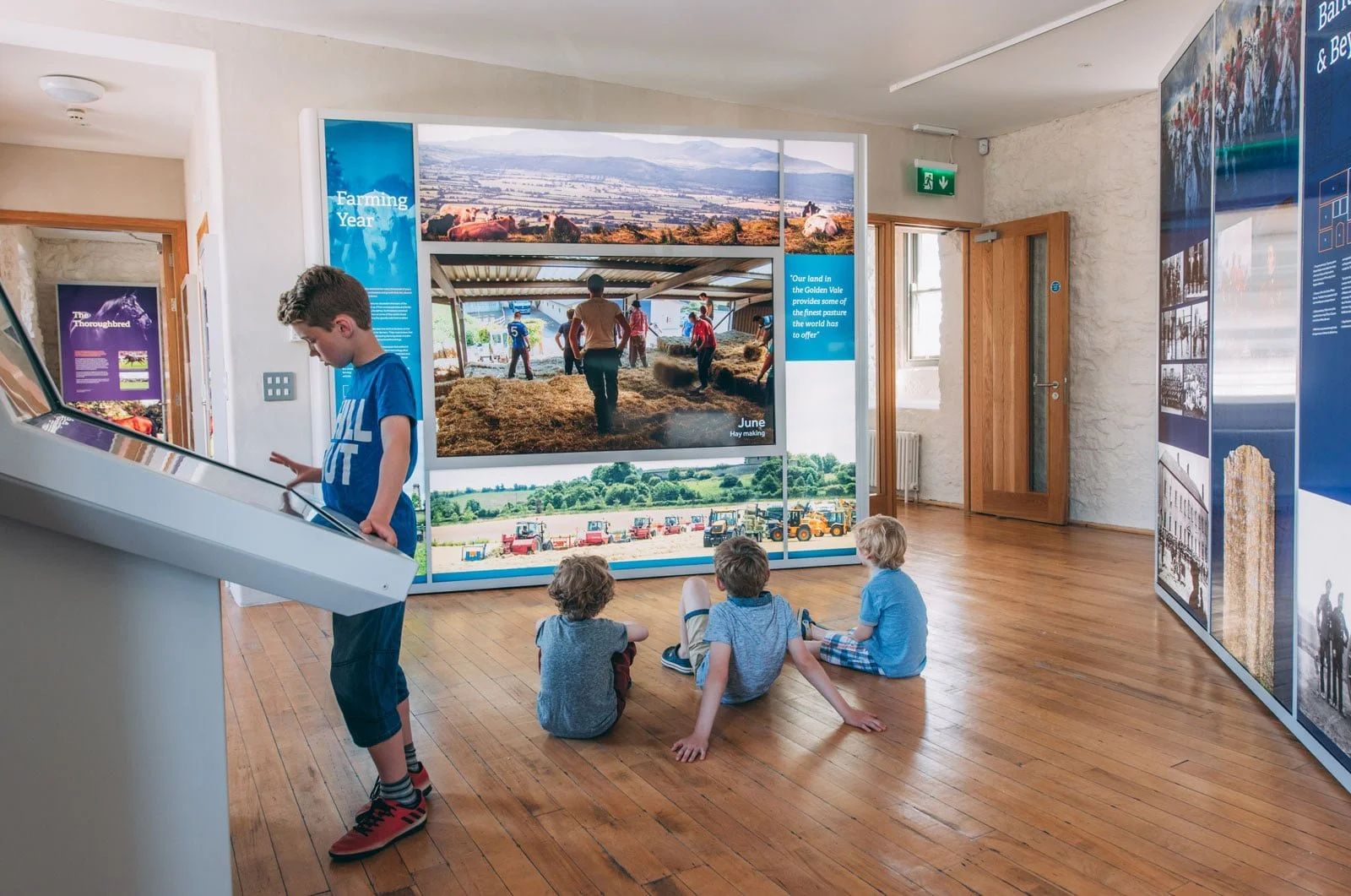 Four children sitting and standing in a museum exhibit room with informational panels and photos of farming activities. One child is standing at an exhibit, while three are sitting on the floor.