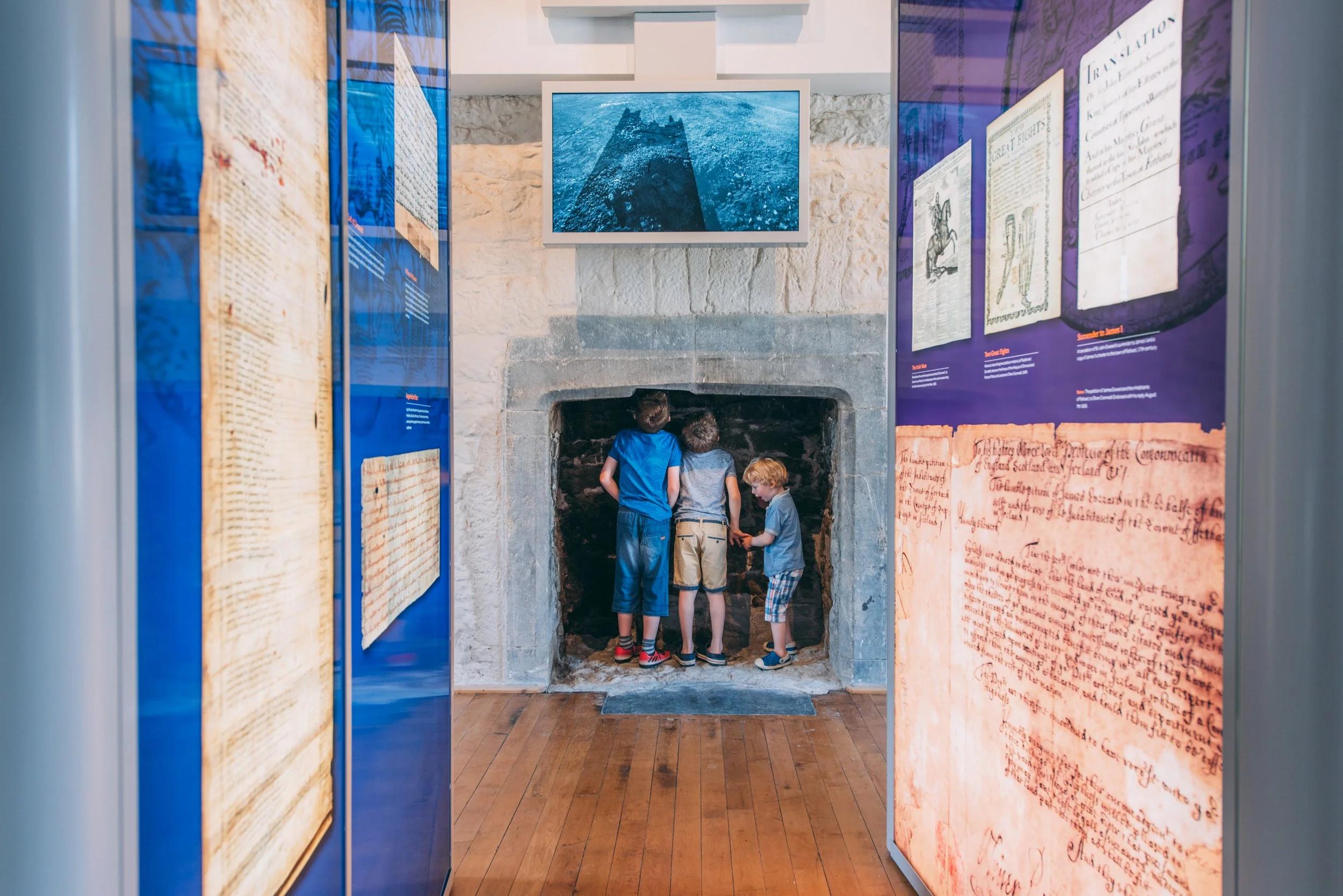 Four children view an old fireplace in a museum exhibit, surrounded by informational panels about historical documents and artifacts.