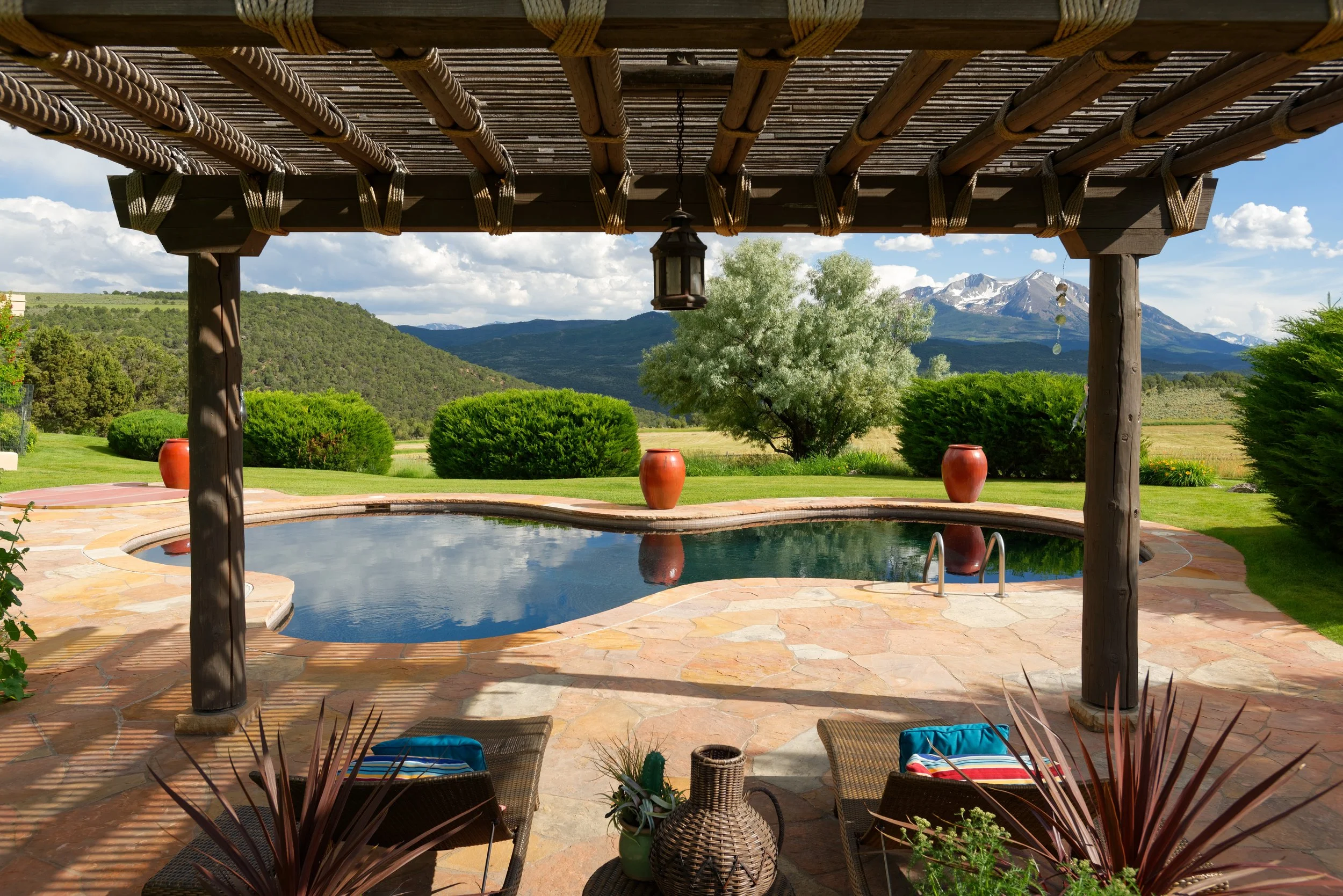 View of a backyard with a kidney-shaped swimming pool, surrounded by greenery, mountains in the distance, and a shade structure with a hanging lantern in the foreground.
