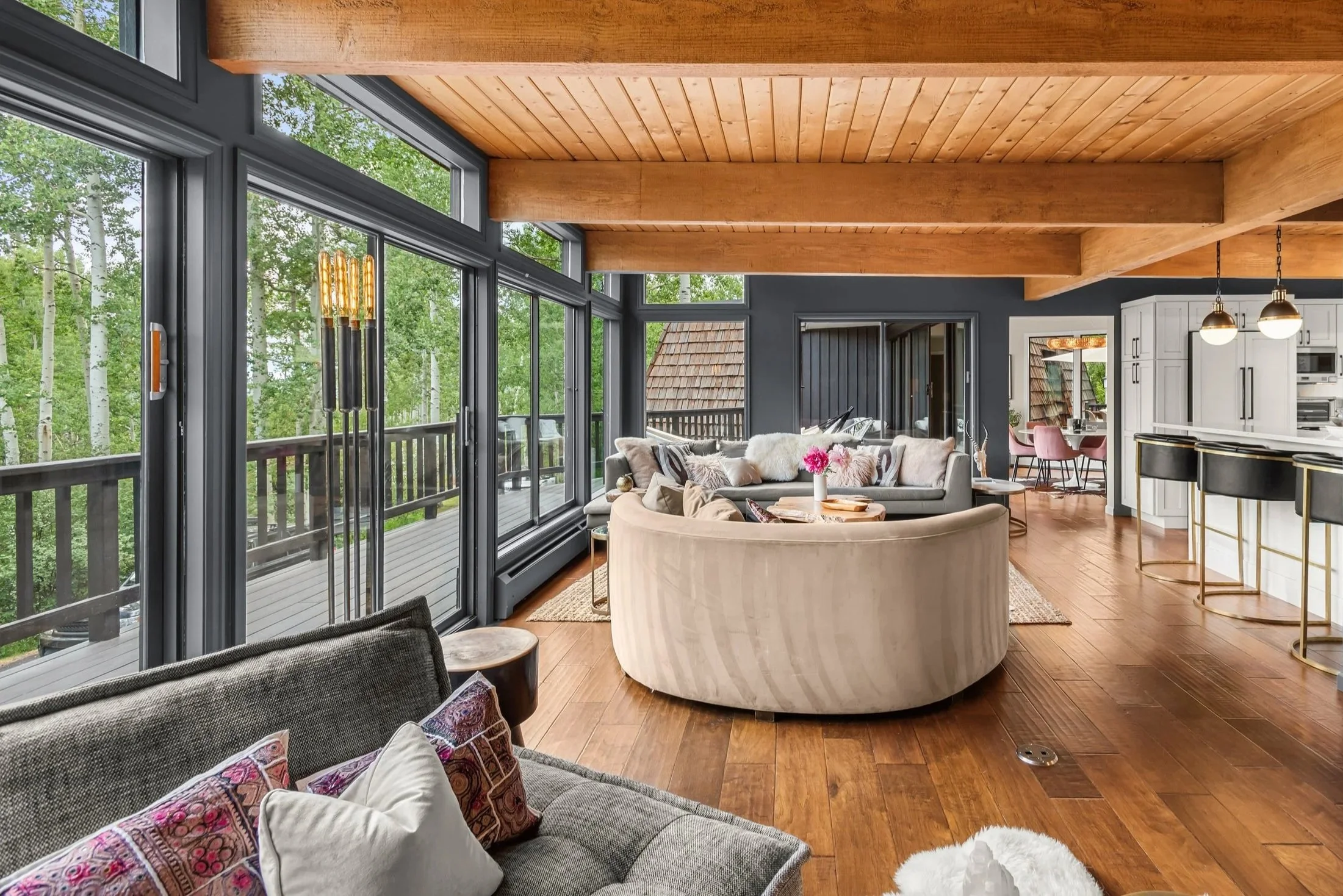 Living room with large glass windows overlooking a forest, modern furniture, a round sofa, a gray couch with pillows, wooden floors, and a kitchen area with bar stools and pendant lights.